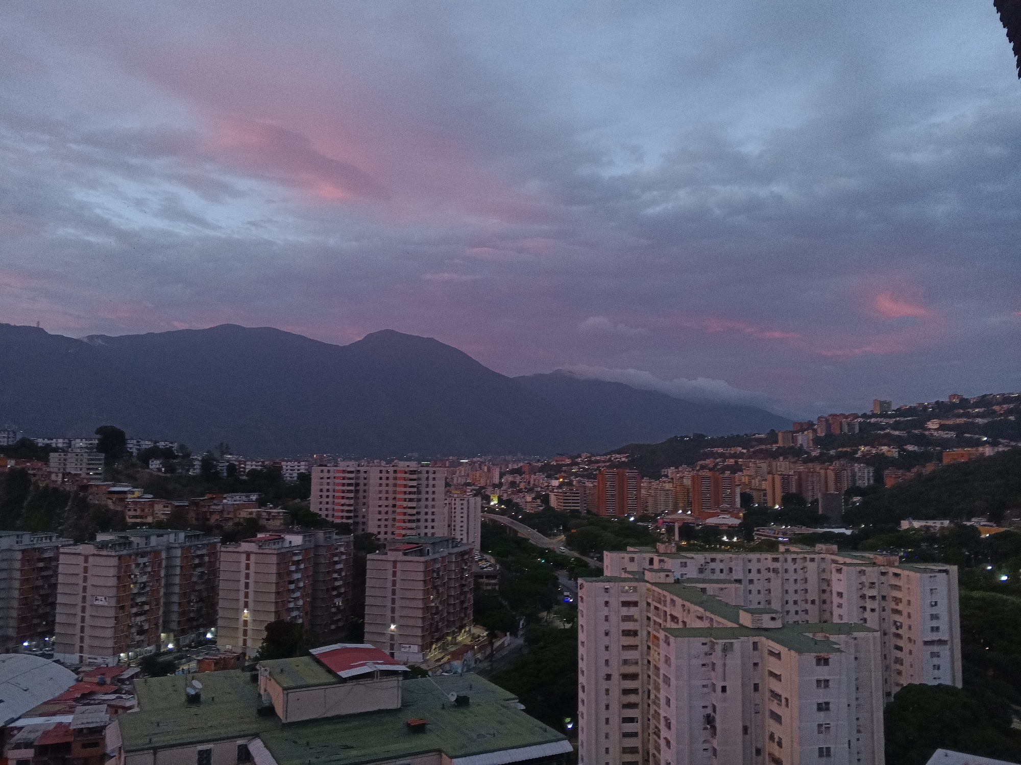 The image captures a panoramic view of Caracas, Venezuela at dusk. In the foreground, densely packed residential buildings with repetitive designs dominate, their colors muted by the fading light. A highway with minimal traffic runs through the middle ground, cutting through a green belt. In the background, a dark mountain range forms a stark contrast to the illuminated city. The sky is a blend of gray and pink hues, suggesting the end of sunset. There are no people visible. The overall environment evokes an urban landscape transitioning from day to night.