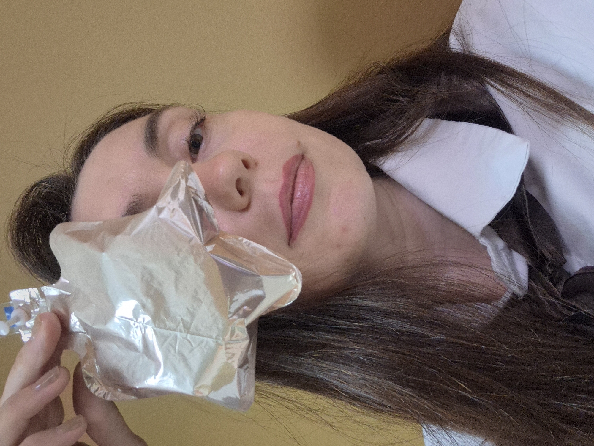A woman with long brown hair, wearing a white collared shirt, is holding a sealed, silvery, translucent bag near her face. The location is indoors, against a yellow wall. The city and specific location within it are unknown.