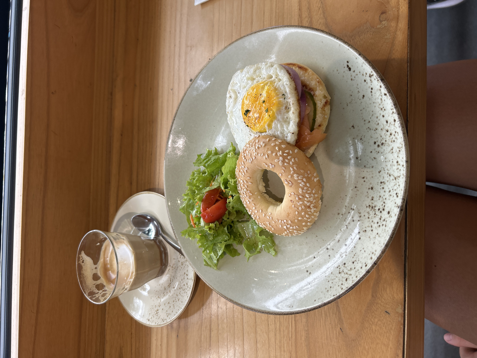 A slightly overhead shot shows a plate of food and a glass of coffee on a wooden table. The plate contains a halved bagel topped with smoked salmon, red onion, and a fried egg. Next to the bagel is a small salad of lettuce and tomato. A sesame seed bagel half is also on the plate. A glass of coffee with a saucer and spoon is on the left side of the frame. The table appears to be wooden. Part of a person's leg is visible on the right side of the frame. The location is assumed to be in Kuta Selatan, Indonesia based on the request.