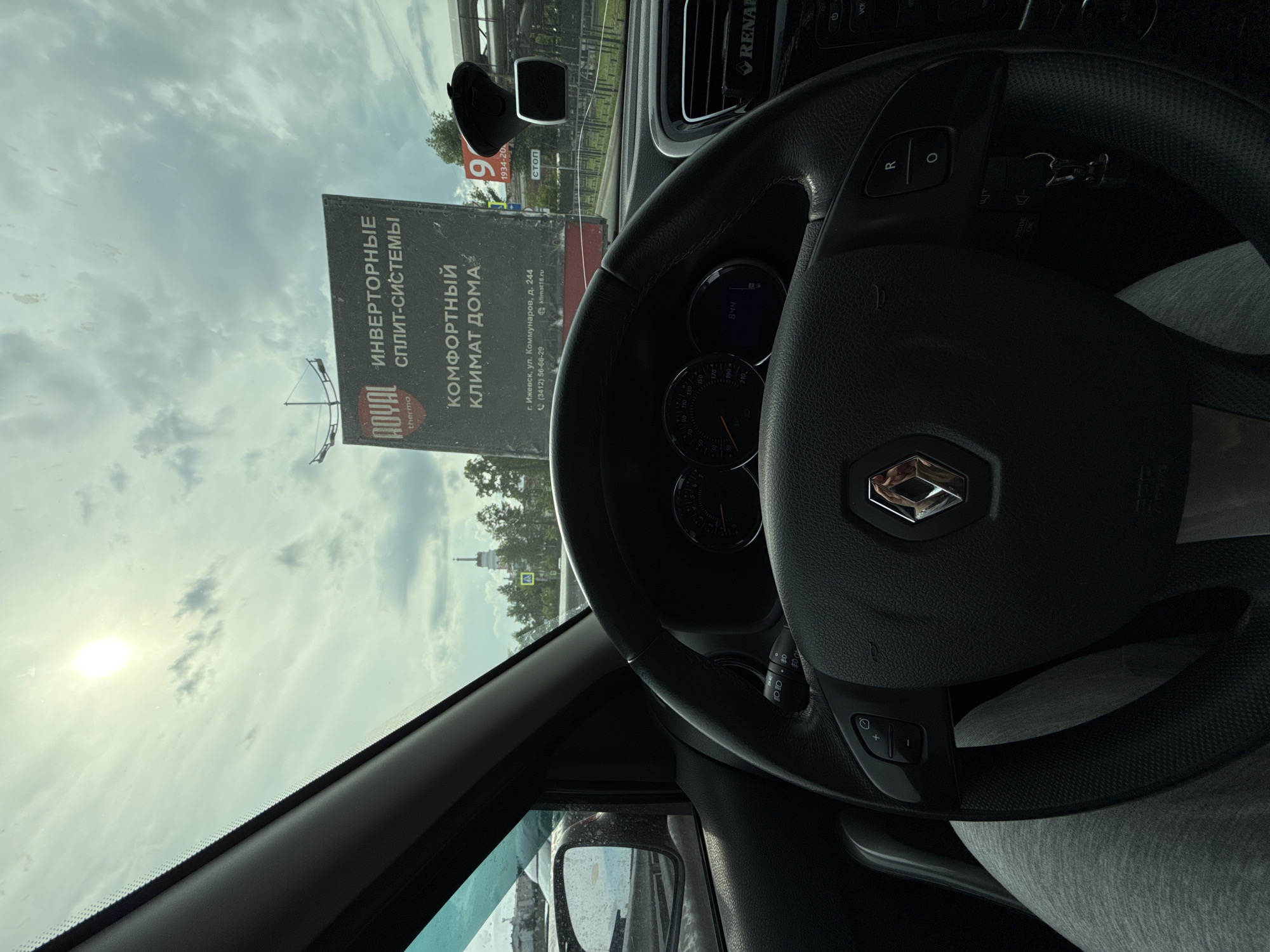 The image captures a first-person perspective from inside a car, looking out through the windshield. A black steering wheel, bearing a silver Renault logo, occupies a significant portion of the right side, partially obscuring the dashboard, instrument cluster showing "844", and a RENAULT branded air vent. The driver's lower body, clad in grey sweatpants, is visible beneath the steering wheel. Controls like "R", a circle/slash, "MODE", and "OK" are visible on the steering wheel and its stalks. A phone mount is attached to the windshield.
Outside the car, a large vertical billboard is prominent against a cloudy but bright sky, with the sun creating a glare in the upper left. The billboard features Russian text, advertising "ИНВЕРТОРНЫЕ СПЛИТ-СИСТЕМЫ" (Inverter Split Systems) and "КОМФОРТНЫЙ КЛИМАТ ДОМА" (Comfortable Home Climate) for the "ROYAL thermo" brand. Contact information provided includes "г. Ижевск, ул. Коммунаров, д. 244" (Izhevsk, Kommunarov Street, 244), a phone number "(3412) 56-66-29", and a website "klimat18.ru".
Further down the street, a small sign with "СТОП" (STOP) is visible. In the distance, beyond trees, the spire of a white building, possibly a church or cathedral, can be seen against the sky. The time of day is clearly daytime, and the weather is partly cloudy. The scene is set in an urban or suburban environment in Izhevsk, Russia.