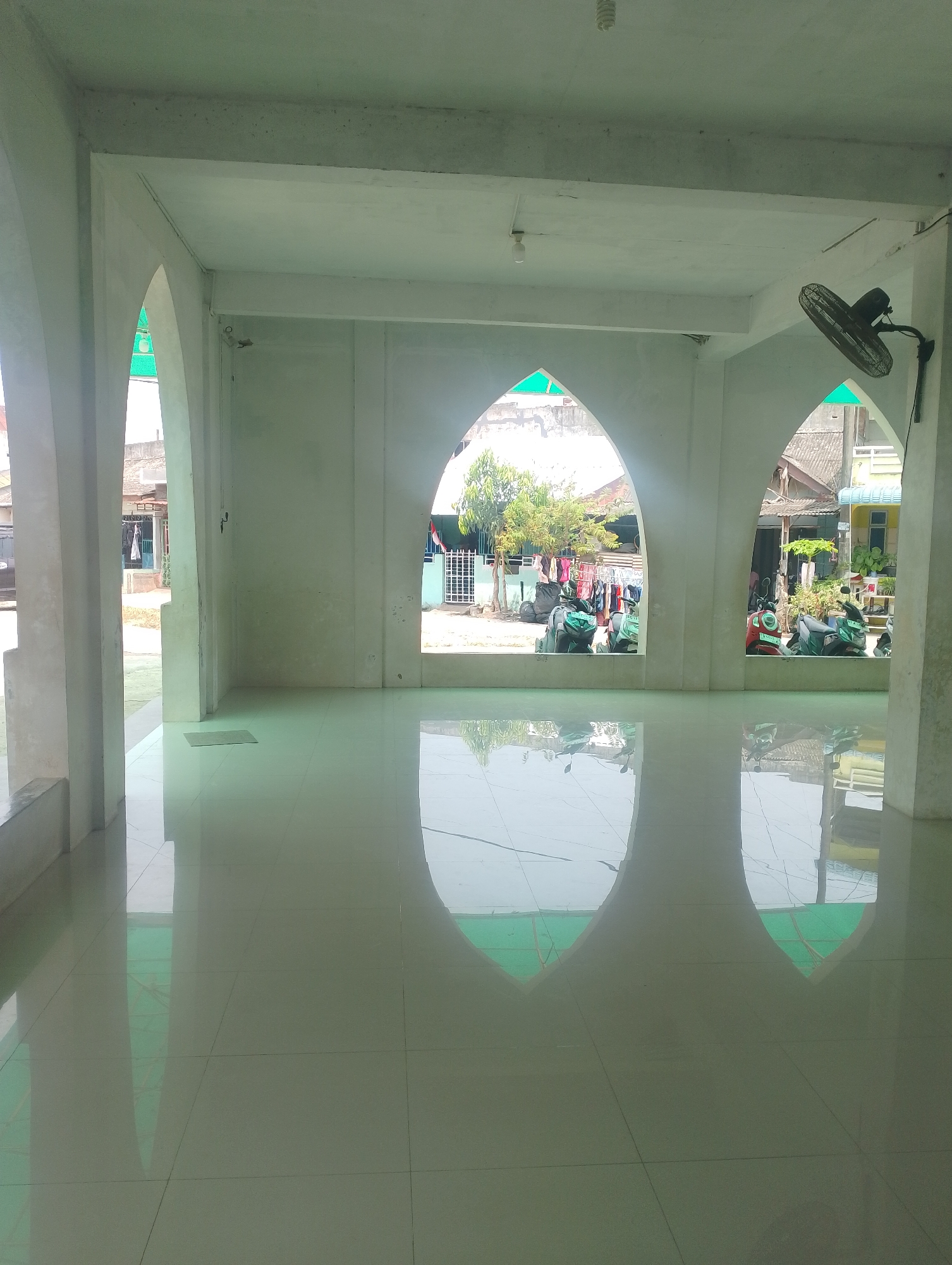 The image depicts the interior of a building, possibly a mosque or community hall, with a highly polished, reflective floor. Sunlight streams in through arched openings, creating reflections on the floor. Outside, a residential area is visible with houses, laundry hanging on a clothesline, and several motorcycles parked. The architecture features white walls and columns, with a black oscillating fan mounted on the wall in the upper right corner. The scene appears to be captured in daylight, likely during a warm season. The location is identified as Batam, Indonesia.