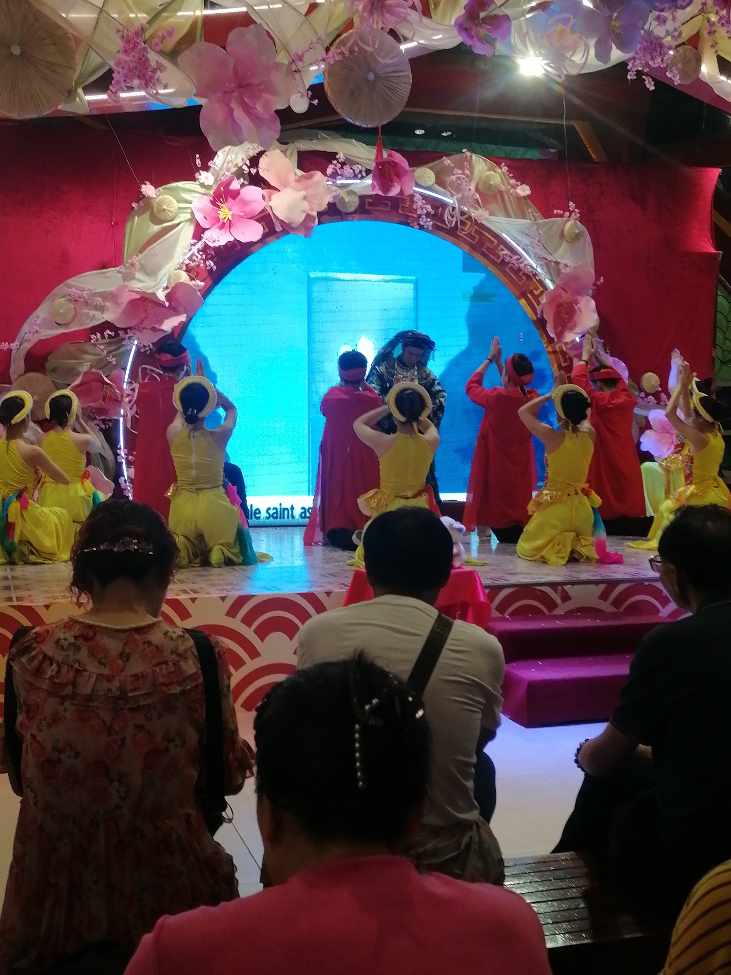 Captured in Hong Gai Ward, Vietnam, the image presents a performance on a stage adorned with a large, floral-decorated archway. Performers in red and yellow costumes are engaged in what appears to be a dance or theatrical act. The audience is visible from the rear, watching the performance. The environment is indoors, likely in a theater or event space.