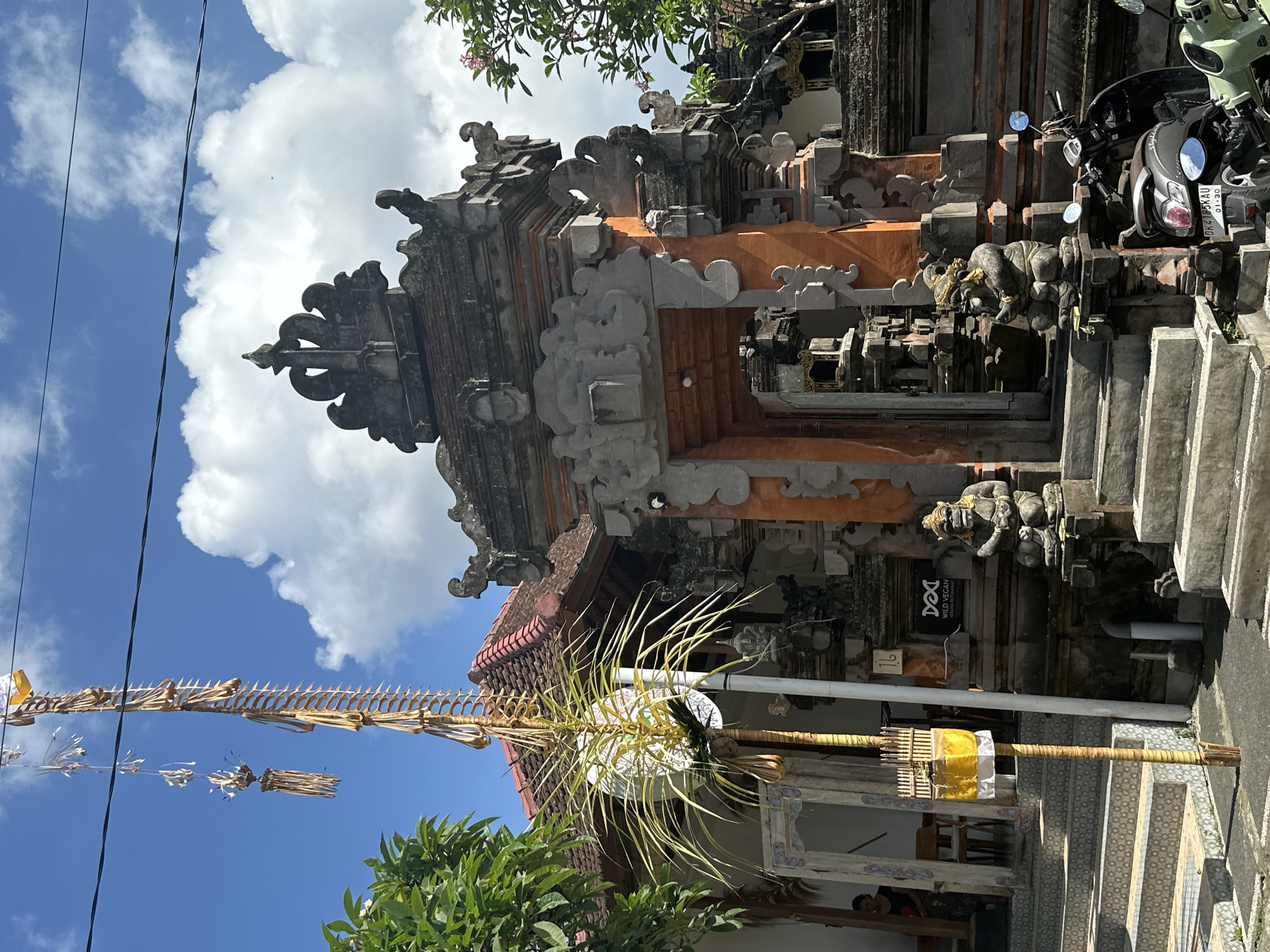 The image depicts the entrance to a building in Ubud, Indonesia, likely a temple or traditional establishment. The architecture features ornate stone carvings and orange-colored stucco. Two guardian statues, resembling monkeys or mythical beings, flank the main entrance. A tall, decorative bamboo pole adorned with leaves and fabric stands prominently in the foreground, suggesting a cultural or religious ceremony. Steps lead up to the entrance, and a scooter is parked to the right. Lush green trees and a bright blue sky with scattered clouds form the background. A sign that reads "WILD VEGAN" is visible on the building's facade.