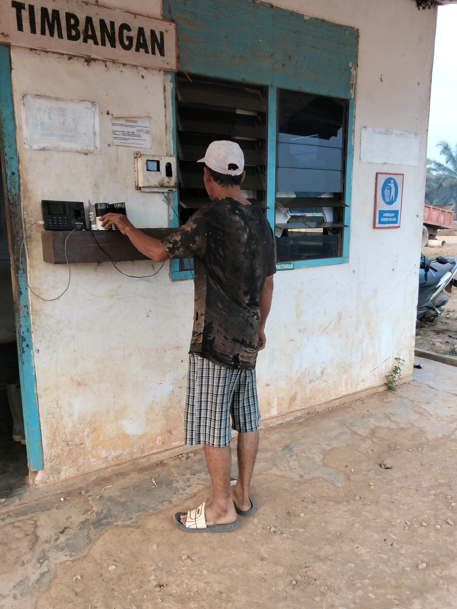 In Teluk Tenggirik, Indonesia, a man wearing a white cap, a dirty black t-shirt, and plaid shorts stands with his back to the camera, interacting with a time clock device mounted on a wooden shelf. The time clock is black and rectangular with a small screen and keypad. To the right of the time clock, on the same shelf, are two cylindrical objects. The man's right hand is extended towards these objects. The scene takes place in front of a weathered, off-white building with blue trim around a window with horizontal louvers. A sign with large black letters reading "TIMBANGAN" is mounted above the building's entrance. On the wall to the left of the window, there is a document affixed, and to the right, a blue and white sign depicting a trash can with a line through it and the text "JAGALAH KEBERSIHAN" (Maintain Cleanliness). The ground is a dirt and concrete mixture. In the background, a motorcycle is parked to the right, and parts of a red truck are visible.