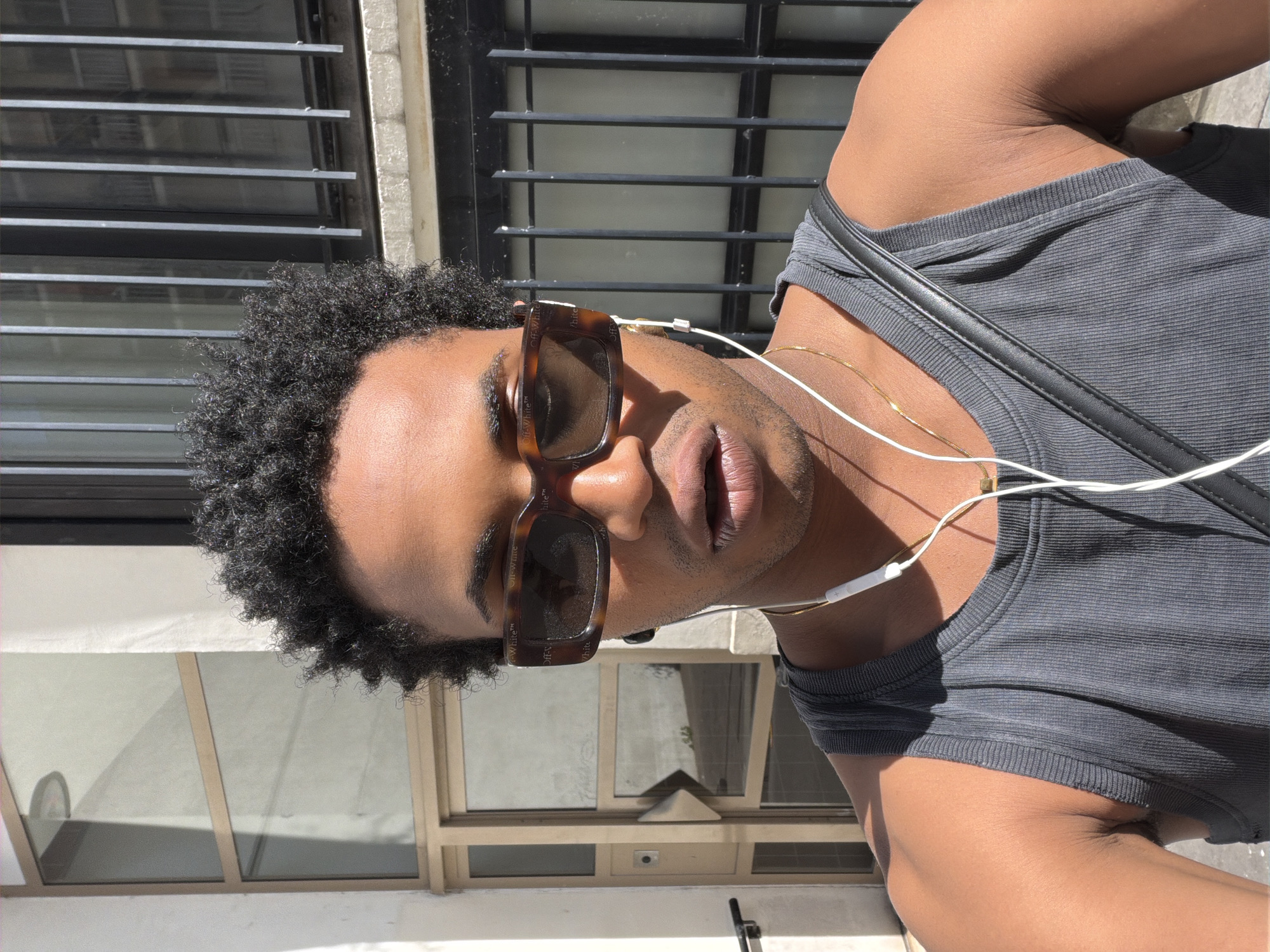 A man is the primary subject of the image, which appears to be a selfie taken in an urban setting. He is facing the camera and wearing sunglasses and wired earbuds. His hair is short and curly. He is wearing a gray tank top and a thin gold chain. The background indicates an outdoor setting, likely a city street, with building facades and windows visible. Based on the architectural style of the buildings in the background, the location could potentially be in Paris, France.