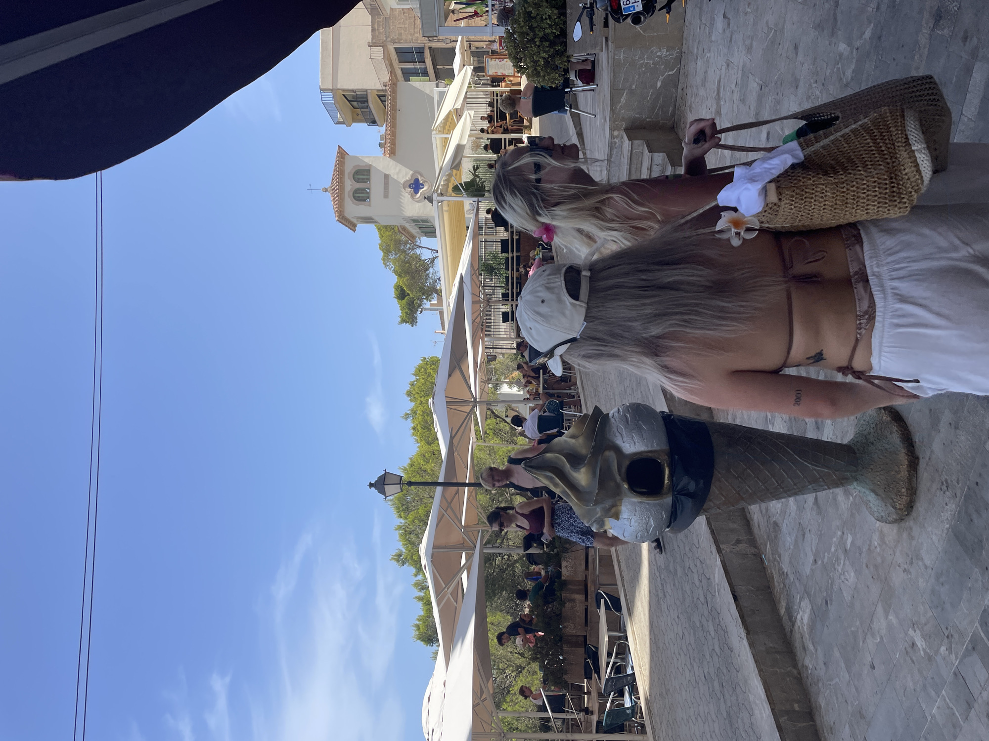 The image depicts a sunny outdoor scene in Santanyí, Spain, likely a tourist area with outdoor dining. In the foreground, two women are seen from behind, walking along a paved walkway. The woman on the left wears a white baseball cap and her blonde hair is visible. The woman on the right has blonde hair adorned with a pink flower, and she is carrying a woven tote bag filled with items.
To the left of the women is a large, metallic sculpture of an ice cream cone with a scoop, which appears to be a public trash receptacle. In the middle ground, several tables and chairs are set up under large, light-colored umbrellas, suggesting a cafe or restaurant. People are seated at some of these tables, engaged in conversation or dining. Behind the umbrellas, lush green trees and foliage are visible, and further in the background, a multi-story building with balconies and a tower-like structure with arched windows can be seen. The sky is bright blue with scattered white clouds. The overall atmosphere suggests a warm, pleasant day in a Mediterranean setting.
