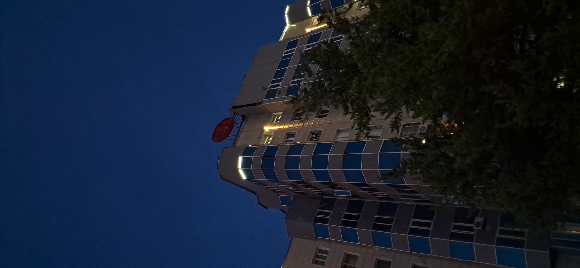 An exterior, low-angle, vertical shot captures the upper section of a modern, multi-story building against a dark blue night sky. The building's facade is a mix of light grey panels and a pattern of blue-tinted glass windows. Some windows are lit from within, creating rectangular reflections. Along the top edge of the building, decorative white lights are visible, illuminating the roofline. A prominent, large, red, circular sign with a dark, ribbed surface is affixed to the rooftop. Green trees, densely leafed, obscure the lower portion of the building, with their branches extending upwards and partially covering some windows. The overall scene suggests an urban setting at dusk or night.