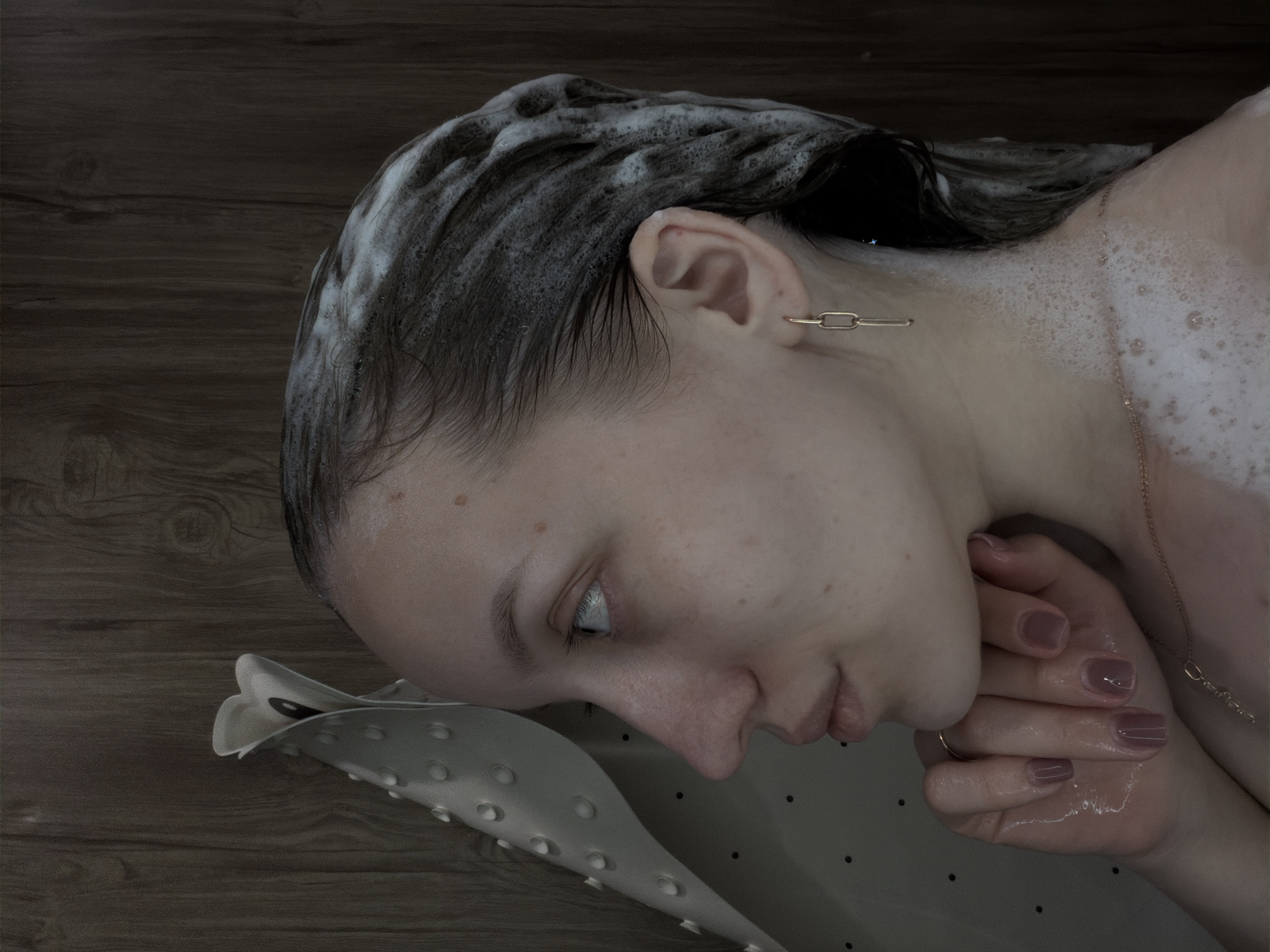 A woman lies in a bathtub, her head resting on a light gray, molded bath pillow. Her dark hair is covered with white soap suds. She is wearing a gold chain earring and a necklace. Her nails are painted a mauve color. She is holding her hands together under her chin. The background appears to be a dark brown wood paneling. The location is Reshetnikovo, Russia.
