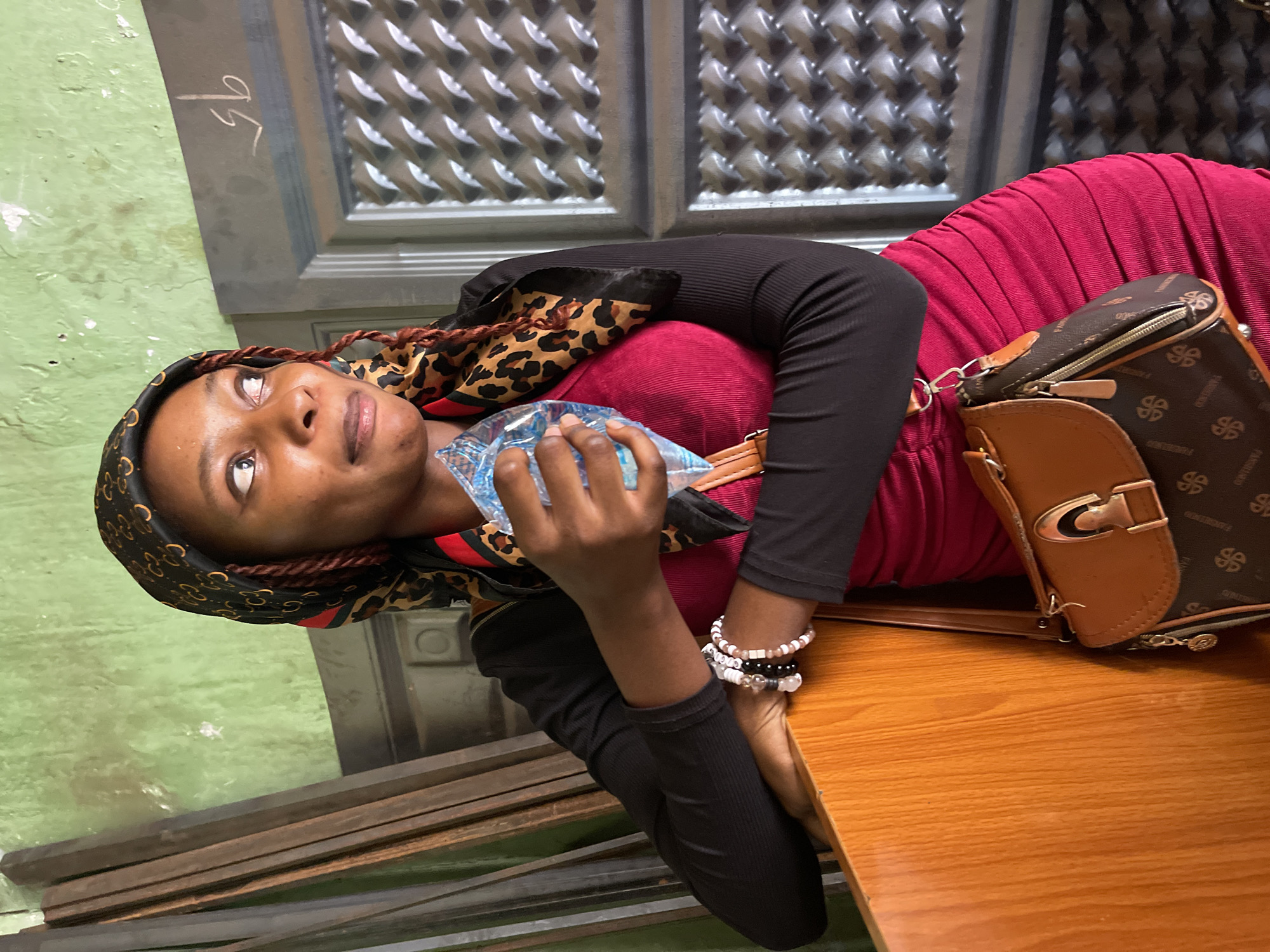 The image shows a young woman lying down in an indoor setting, looking upwards with a relaxed, slightly thoughtful expression. She is dressed in a black long-sleeved top, a vibrant red ribbed or pleated skirt, and a patterned headscarf featuring leopard print alongside black and red details, revealing reddish-brown braided hair.
In her left hand, she holds a small, clear plastic packet, possibly containing water or ice. Her left wrist is adorned with a stack of beaded bracelets in white, black, and pink colors. A brown and dark brown patterned shoulder bag with gold hardware rests on a light brown wooden surface, likely a table or bench, beside her. The bag's pattern includes what appears to be text, possibly "PARISLAND" and "FENDIBAG".
The background features a light green wall or floor on the left, with several dark, rectangular objects resembling metal bars lying on the ground. To her right and above, there is a dark gray structure, possibly a wall or door, which has a textured, patterned surface and the white numbers "95" visibly written on it. The diffused lighting suggests an indoor environment, perhaps a break area or waiting room in Zaria, Nigeria.