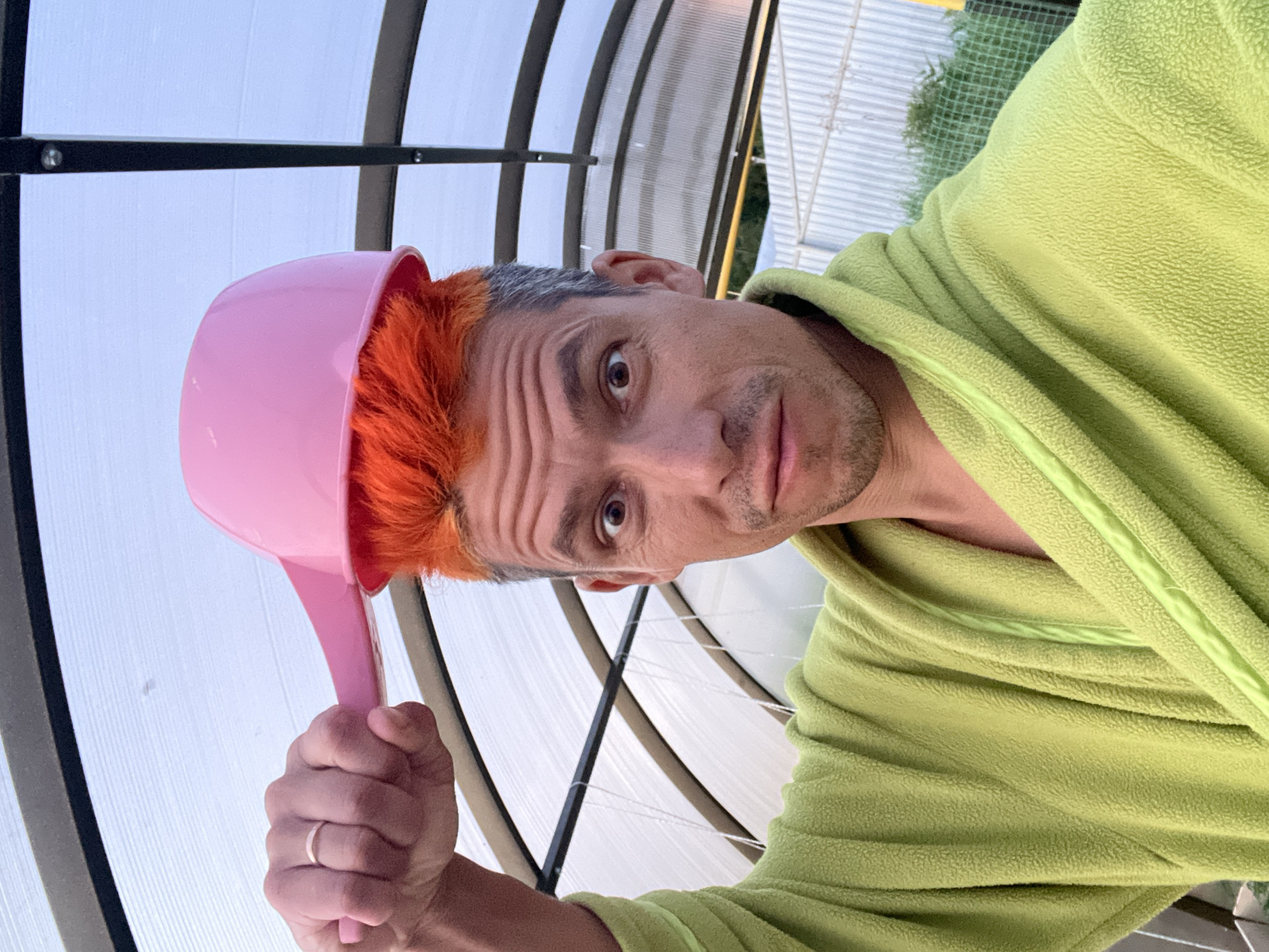 A close-up, slightly distorted selfie captures a man from a low angle, looking directly at the camera with a surprised or wide-eyed expression. His left eyebrow is notably furrowed, creating multiple horizontal lines on his forehead, while his right eyebrow is slightly raised. He has prominent orange hair on the top of his head, contrasting with gray hair at the temples, and a short beard or stubble on his chin and jawline.
He is wearing a lime green, textured fleece robe or jacket. On his head, he wears a bright pink plastic basin or bowl, which he also appears to be holding up with his right hand, clenched into a fist, with a light-colored ring visible on his ring finger. The pose and attire suggest a playful or humorous act.
The background reveals a translucent, possibly corrugated or ribbed, arched roof structure, typical of a greenhouse, conservatory, or enclosed patio. Black structural framing is visible throughout the background. In some areas, green netting and hints of outdoor foliage can be seen beyond the translucent panels. The lighting is bright and even, indicating daytime. No visible text is present in the image. The location is Chaikovsky, Russia.