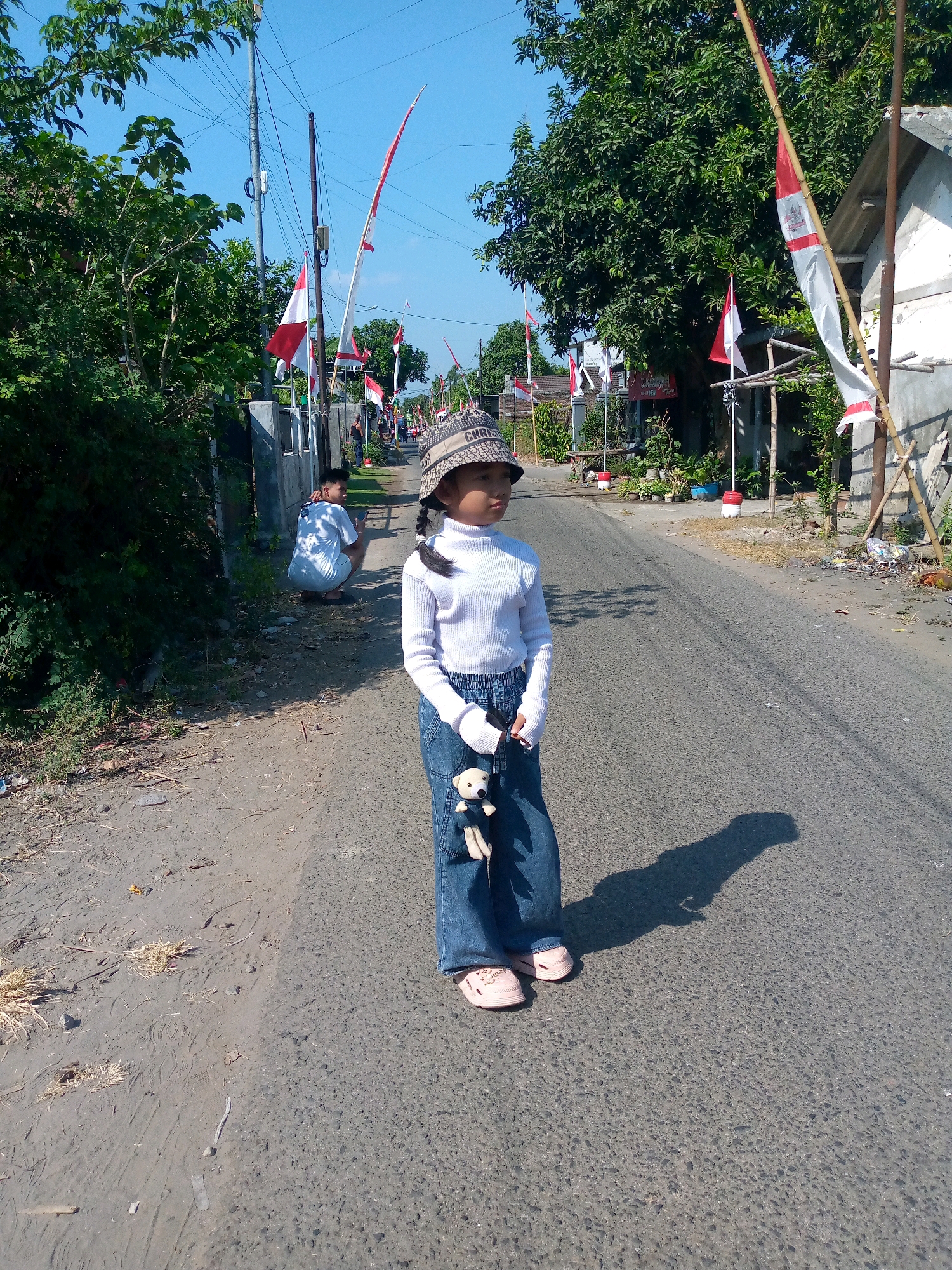 A young girl, wearing a white turtleneck sweater, blue jeans, a patterned bucket hat, and pink slip-on shoes, stands in the middle of a paved road in the village of Sumberwono, Indonesia. She is holding a small stuffed bear. The road is lined with trees and houses, and several Indonesian flags are visible, suggesting a celebration or national holiday. A young man is crouched on the side of the road in the background. The sky is clear and blue, indicating a sunny day.