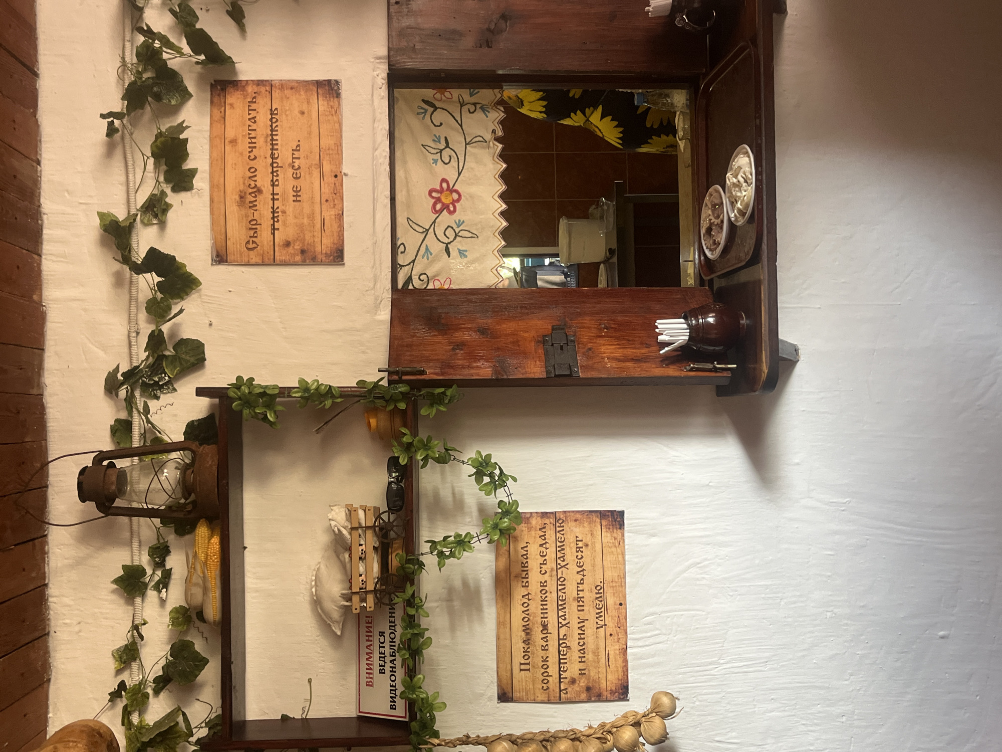 The image depicts a rustic interior scene in Taman', Russia, possibly a restaurant or cafe, characterized by white textured walls and wooden elements. In the upper section, a vintage kerosene lantern hangs above a shelf holding two ears of corn. To the right, a wooden sign with Russian text reads, "Cheese and butter to count, and the dumplings are not there." Another wooden sign below a shelf on the left displays Russian text that translates to "ATTENTION! VIDEO SURVEILLANCE IS BEING CONDUCTED." Below this sign, a small cart holds what appears to be white fabric and sunglasses. To the left, a string of dried garlic hangs down. The central right portion of the image features a wooden cabinet with an open window revealing a glimpse of the interior, including a white bucket and a person wearing a dress with a sunflower pattern. A shelf below this window holds a dark tray with two plates of food, likely dumplings, and a small dark ceramic container filled with white utensils. A decorative vine with green leaves runs across the wall, connecting various elements of the scene.