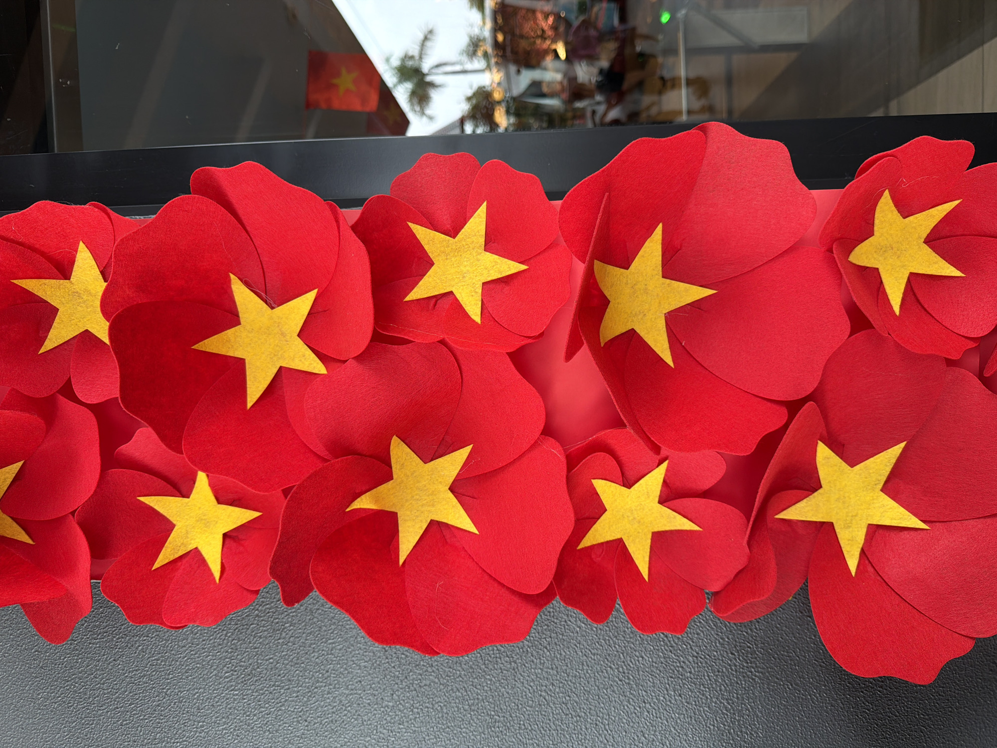 A vertical shot shows a floral decoration on the left side of the frame, composed of large red flowers with yellow stars in their centers. The decoration appears to be made of fabric. The background on the right is a glass window, reflecting the outdoors. Through the glass, one can see palm trees, a street, and people walking by. The reflection also shows a Vietnamese flag with a yellow star on a red background. The overall scene suggests an outdoor setting, possibly a shop or restaurant in Đà Nẵng, Vietnam, decorated for a national holiday or event.