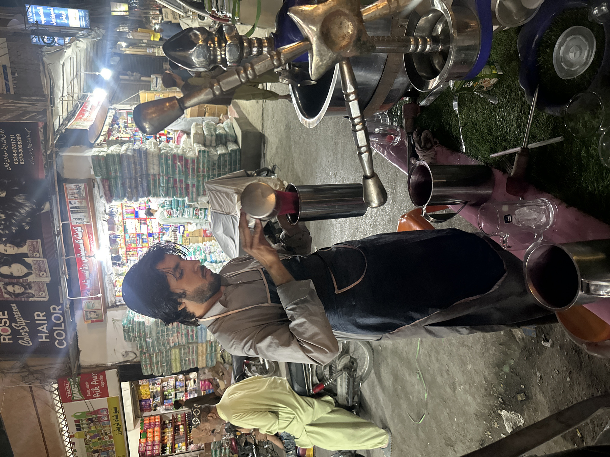 The image shows a street scene in Quetta City Tehsil, Pakistan. In the foreground, a man in a grey shirt and dark apron is pouring liquid from a metal pitcher into a metal container. Several metal vessels are nearby on an artificial lawn mat. Behind him, another man in a yellow shirt and patterned hat is visible. The backdrop features various shops, including one with a "Rose Color Supreme HAIR COLOR" sign and another with "Tapal Dane" branding. Aisles of goods are visible in the stores. The street is bustling, with stacked goods near the shops. Signage includes phone numbers and shop names in Urdu.