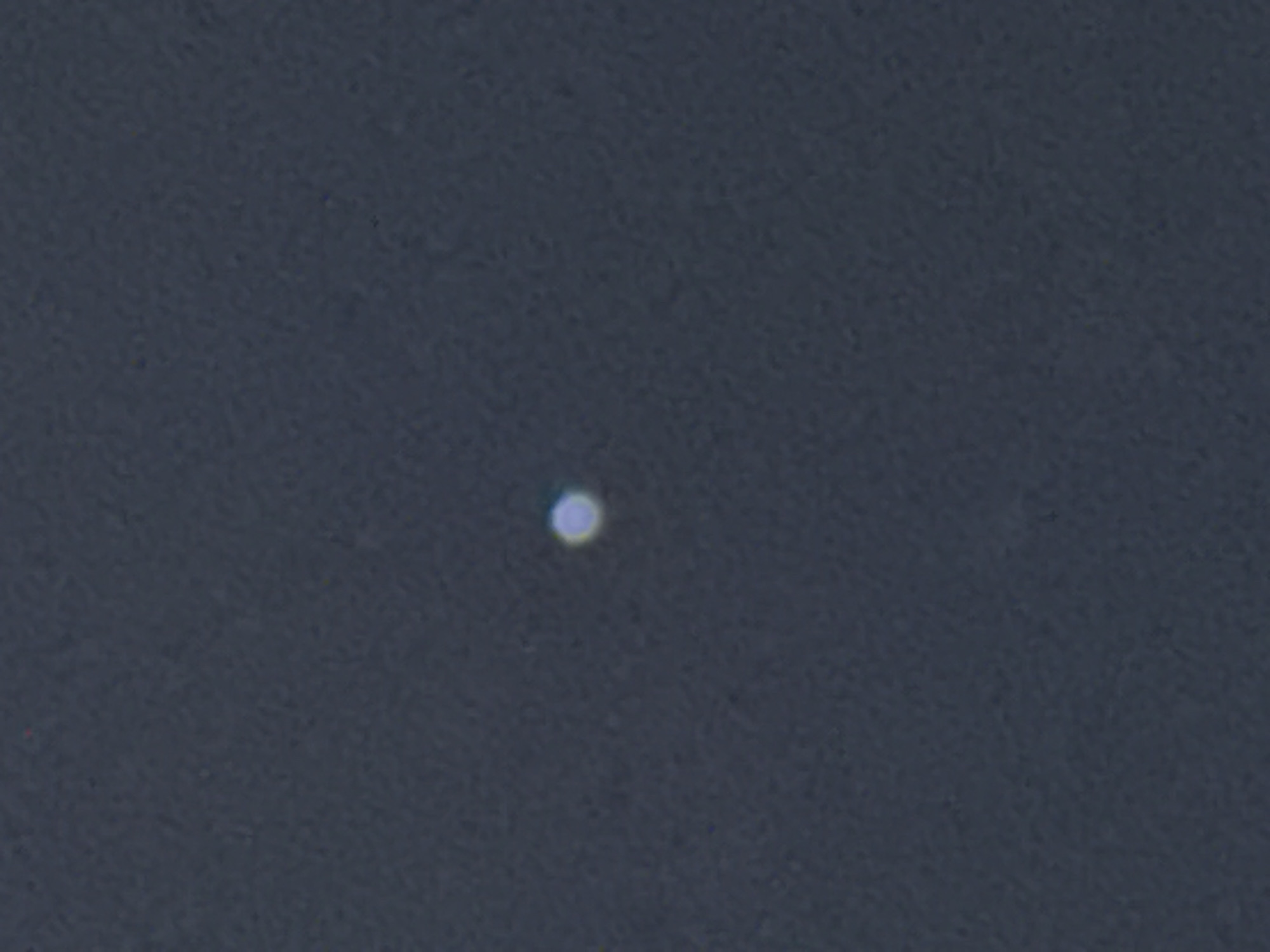 The image shows a dark, out-of-focus background that appears to be the night sky. In the center of the frame, there is a small, bright, circular object with a bluish-white glow and a faint halo. There are no discernible features to indicate its origin or nature, nor any elements that would suggest a specific location within Tbilisi, Georgia.