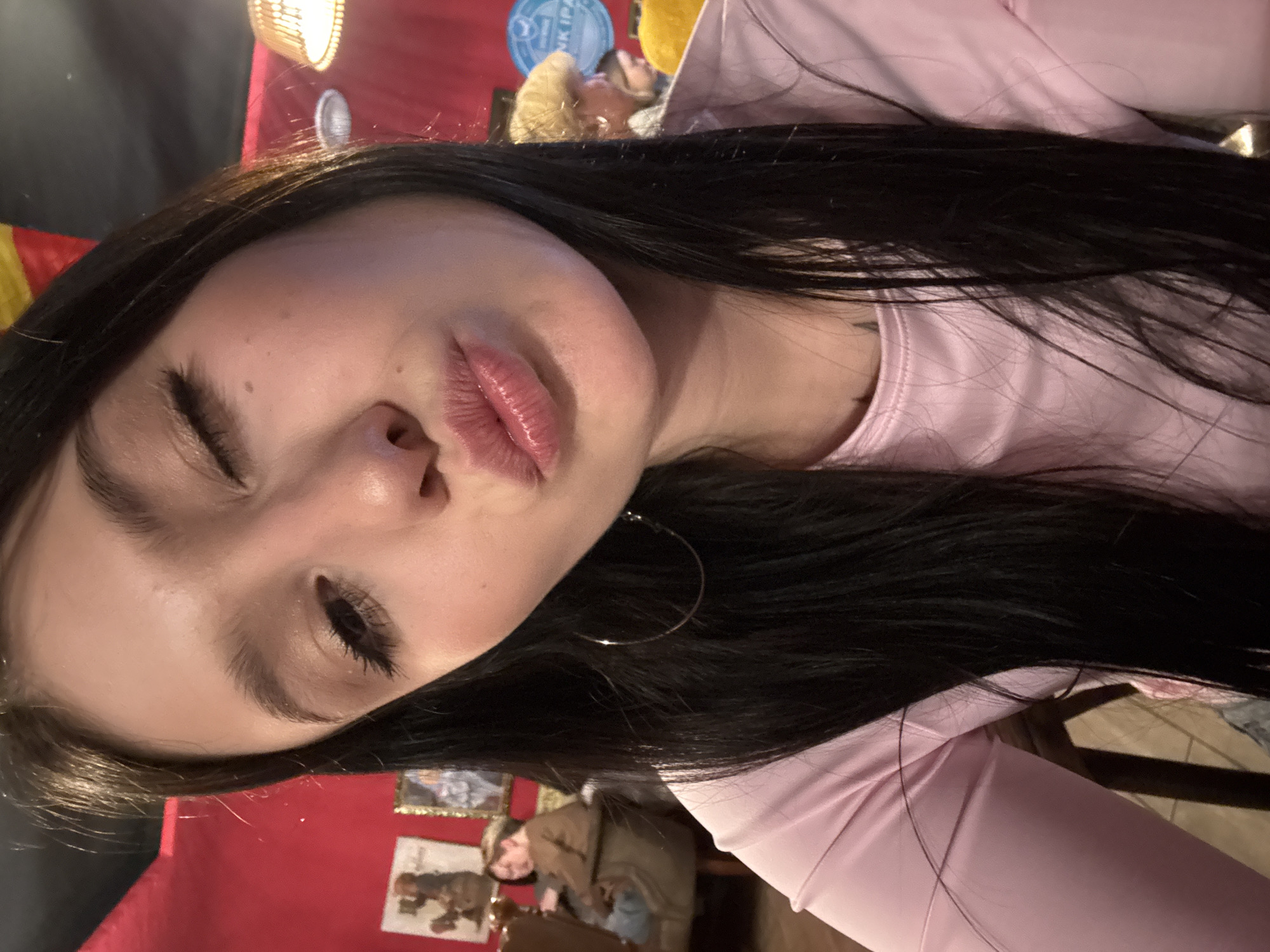 A female with long, dark hair and a light pink long-sleeved top is depicted in a close-up selfie. She is winking with her right eye, while her left eye is slightly open, and her lips are puckered in a kiss gesture. A silver hoop earring is visible on her left ear, and a dark tattoo is partially visible on her upper chest/shoulder area.
The background is an indoor establishment, likely a bar or restaurant, characterized by predominantly red walls and warm, dim lighting. A wicker-style pendant lamp illuminates the scene from the upper right. On the left wall, framed artworks or posters are visible, one featuring a figure in brown tones, and another with a golden frame. On the right wall, a circular blue sign partially displays "NK IPA" in white text. Other patrons are seated in the background in brown upholstered chairs. One individual with brown hair and a brown top is visible on the left, looking downwards, and another person with light-colored hair is partially visible on the right. The specific location within the city of Chaikovsky, Russia, is not identifiable from the visual information presented.