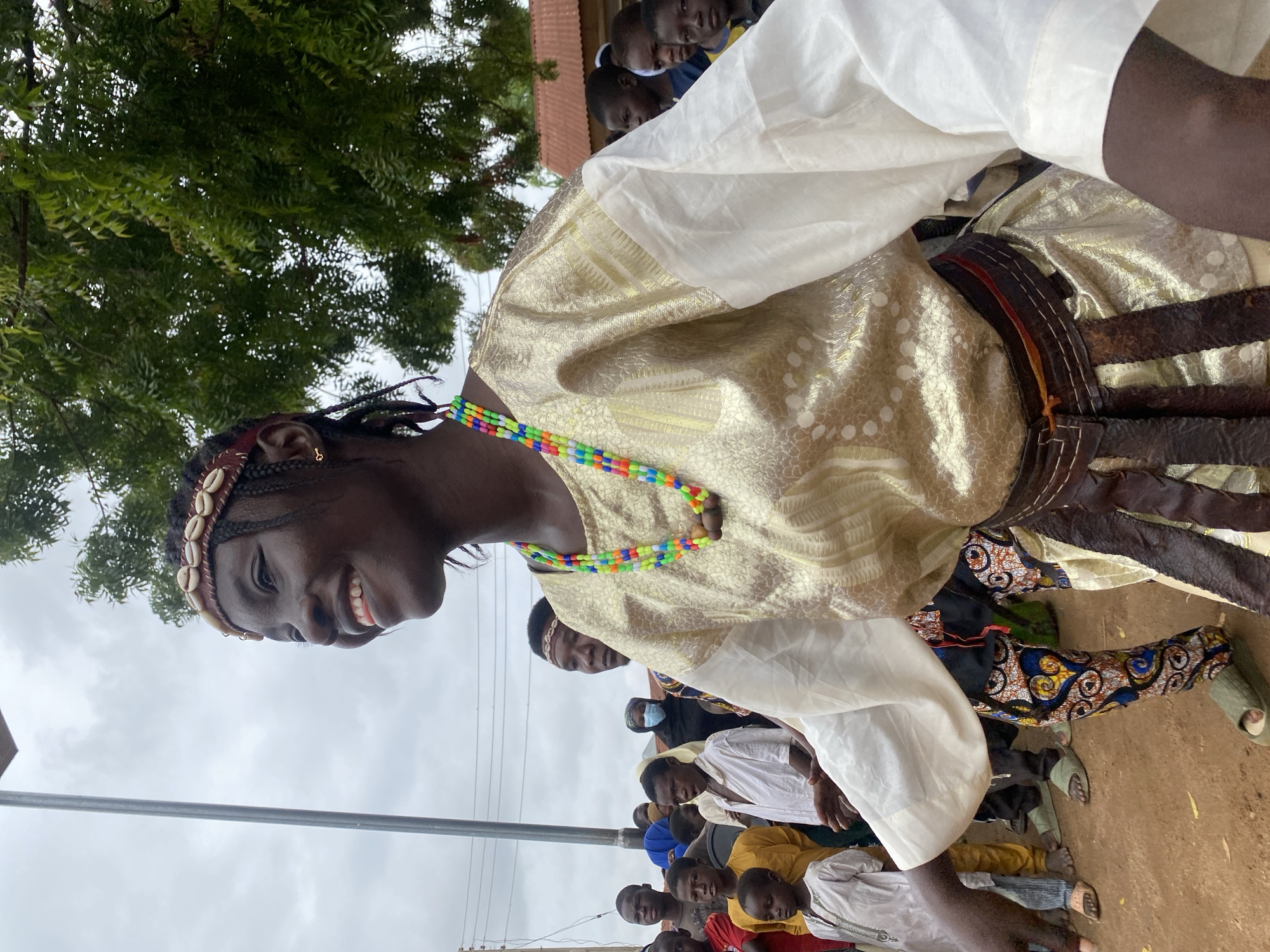 A young Black woman is smiling and looking to her left in a medium close-up shot. She is outdoors in a public space in Rigachikun, Nigeria, with a crowd of people and trees in the background. She has dark skin, braided hair, and is wearing a gold and white patterned robe with a dark leather belt adorned with fringes. A colorful beaded necklace hangs around her neck, and she wears a headband decorated with shells. Several children and adults are visible behind her, some looking towards the camera and others engaged in the event. The ground appears to be dirt, and the sky is overcast. The scene suggests a cultural celebration or gathering.
