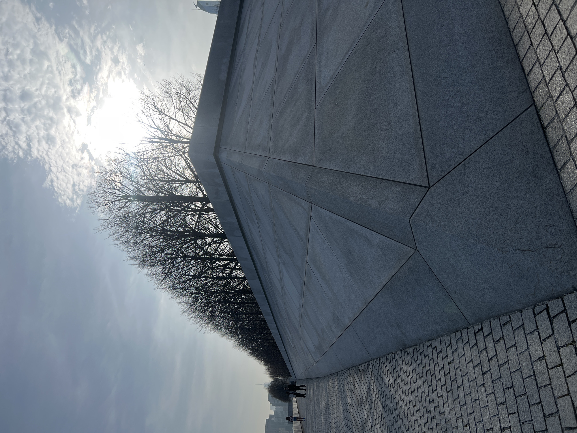 The image depicts a long, gray, textured wall with geometric panels, adjacent to a paved brick walkway. A row of bare trees lines the top of the wall, partially obscuring the bright sun and cloudy sky. In the distance, a few people walk along the walkway, and the faint skyline of New York City is visible. The wall and walkway suggest a public space, possibly a memorial or park in New York City.