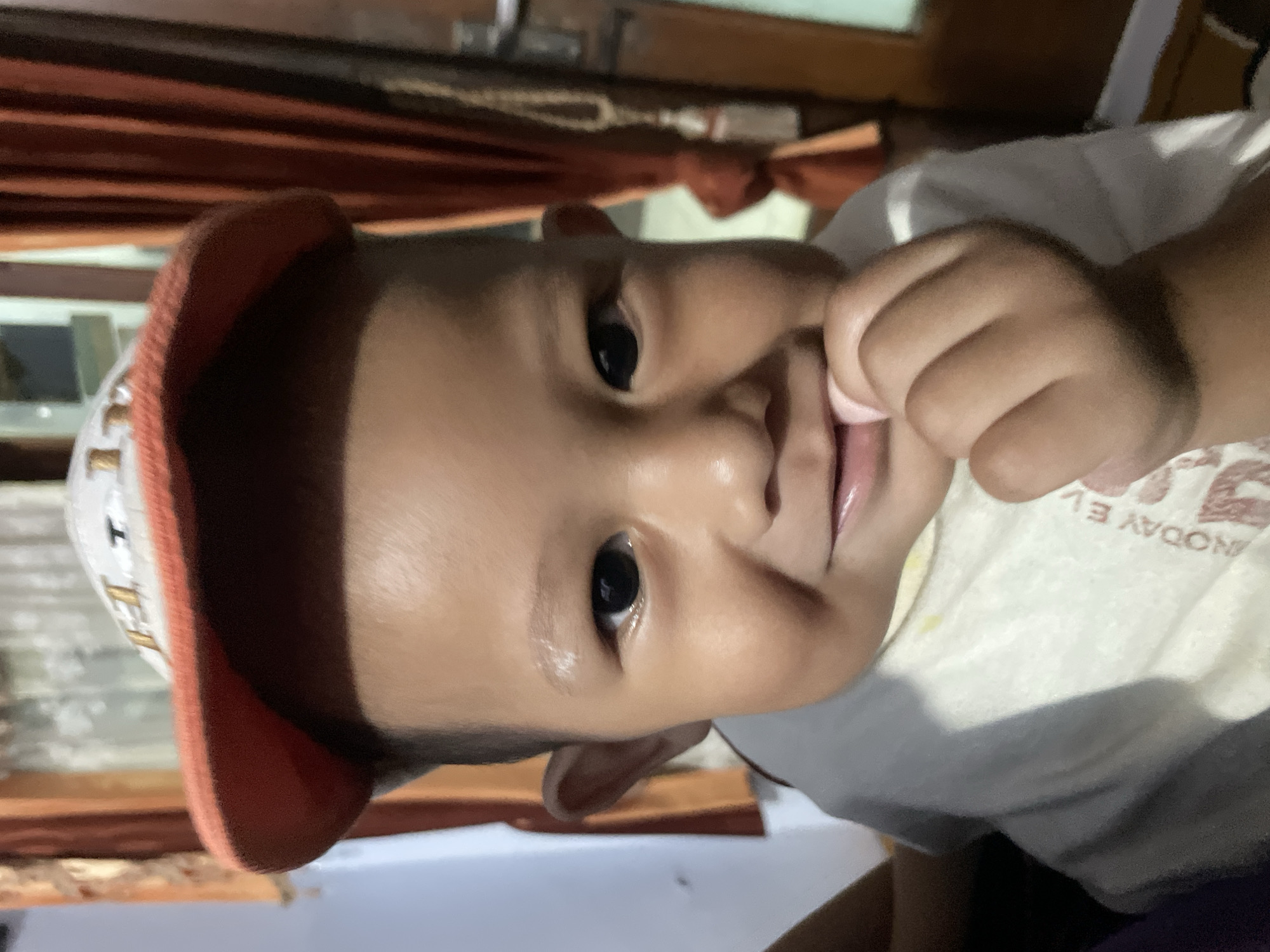 A close-up, eye-level medium shot shows a young child, likely a toddler, wearing a white t-shirt and an orange-brimmed baseball cap. The child is looking directly at the camera with dark eyes and a slight smile, with their right hand brought up to their mouth. The background is blurred, showing orange curtains and a wall, suggesting an indoor setting, possibly a residence in Parakan, Indonesia. The lighting is dim, with some natural light coming from the side.