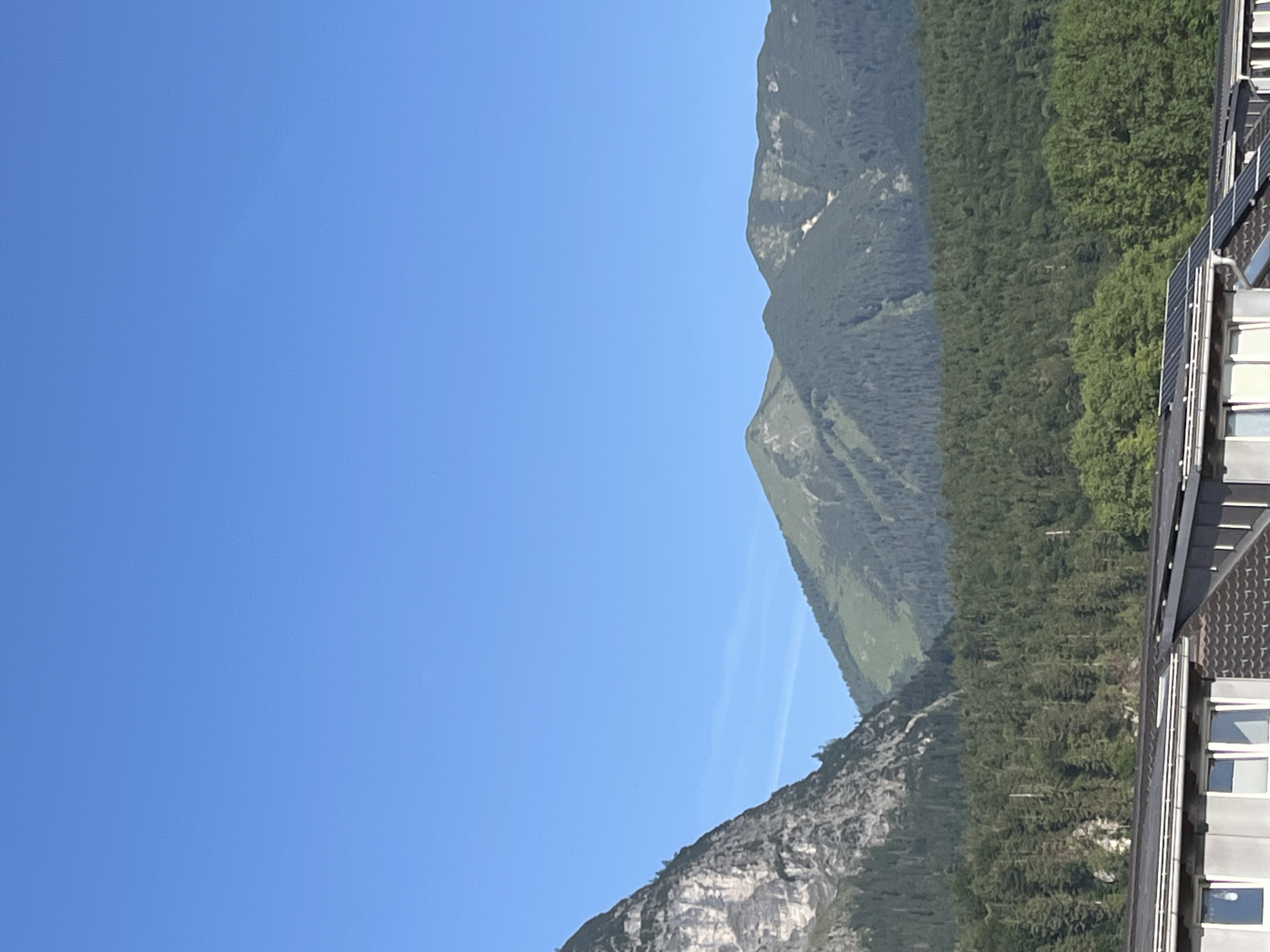 The image shows a clear blue sky over a mountainous landscape. In the foreground, the roof of a building with solar panels is visible. Beyond the building, a dense forest covers the slopes of the mountains. The mountains themselves are rugged and steep, with some patches of green vegetation and rocky outcrops. The overall scene is peaceful and scenic, suggesting a location in a mountainous region such as Garmisch-Partenkirchen, Germany.