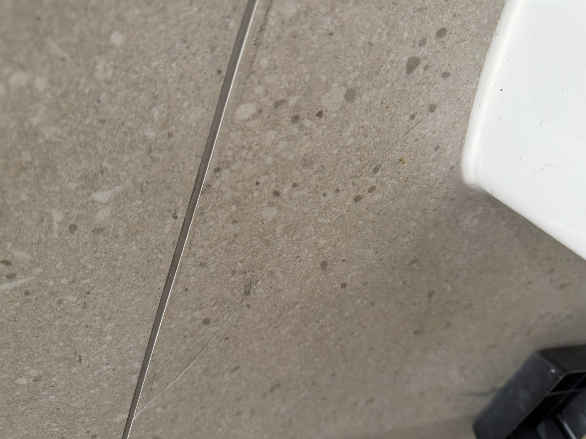 This is a close-up, eye-level, interior shot of a light brown speckled tile wall, possibly in a bathroom or kitchen, in Jerusalem, Israel. A thin, metallic strip runs horizontally across the image, separating two tiles. The tile surface appears slightly textured and has small, darker speckles and some lighter splotches. There are a few faint, thin scratches visible on the surface of the lower tile. In the bottom left corner, a black plastic object, likely a holder or bracket, is partially visible. In the bottom right corner, the curved edge of a white object, possibly a sink or toilet bowl, is in view. The lighting is diffuse, suggesting ambient indoor light.
