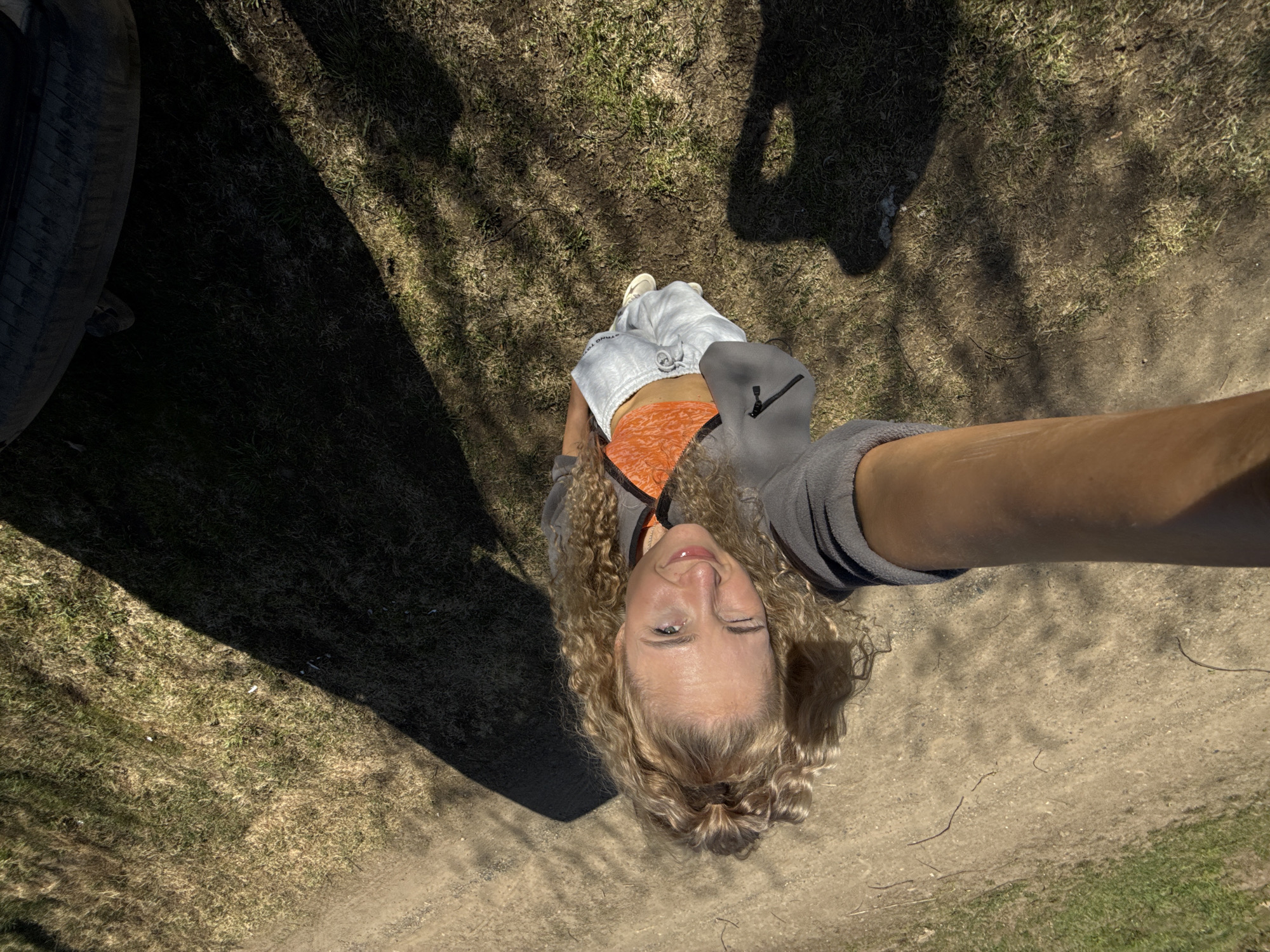 A woman with blond curly hair winks at the camera, holding her arm outstretched to take a selfie. She wears a gray jacket, an orange top, and gray sweatpants, and white sneakers. The background consists of a dirt and grass area with shadows suggesting trees overhead and a tire of a car to the left. The location is inferred, and no elements within the image confirm Rosstani, Russia.
