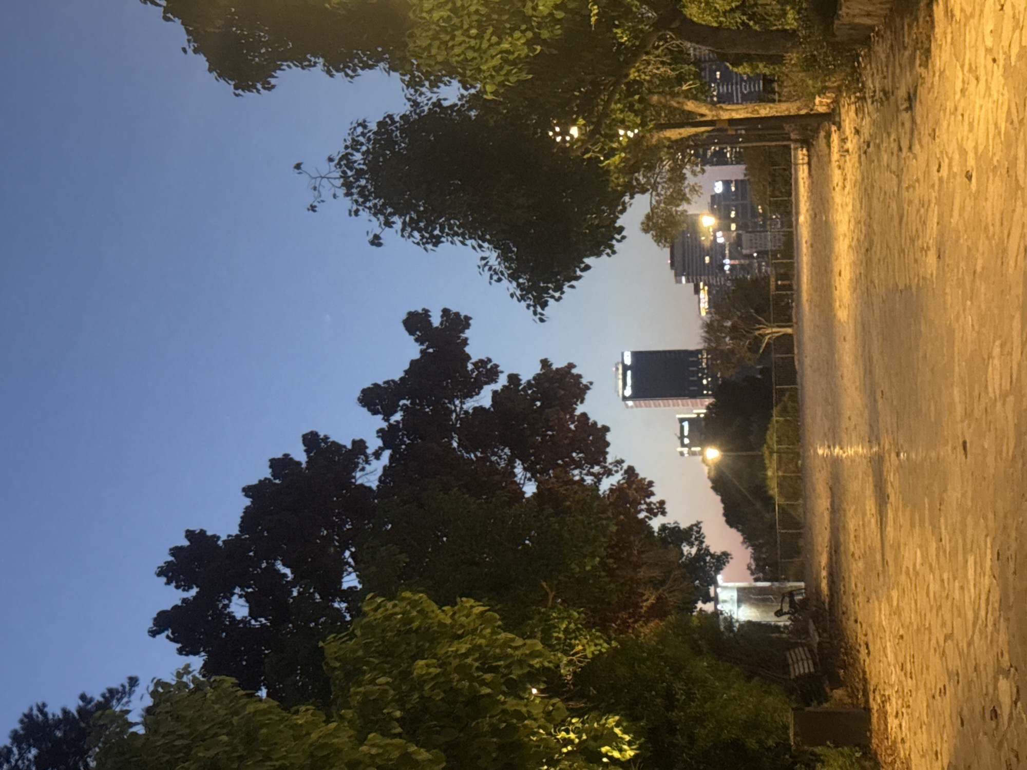 The image depicts a park pathway in Ramat Gan, Israel, at dusk. A cobblestone path, illuminated by artificial lights, stretches into the distance, flanked by trees on either side. Beyond the trees, a city skyline with illuminated buildings is visible. In the foreground, on the left, a park bench and a trash bin are present. The sky is a deep blue, transitioning to a lighter hue near the horizon, indicating the end of daylight. The overall scene is serene, with a mix of natural elements and urban structures.