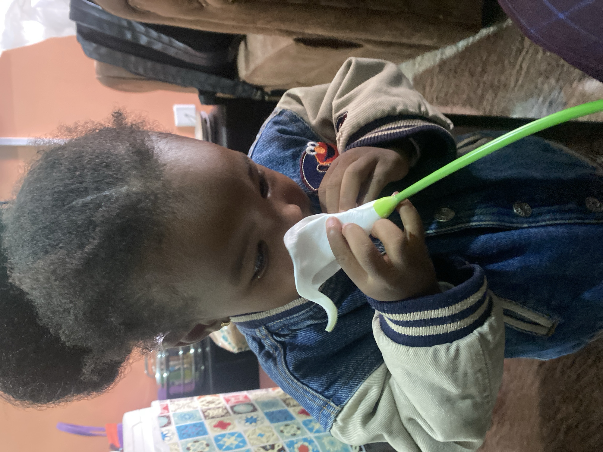 A close-up shows a young child wearing a denim jacket with beige sleeves and striped cuffs. The child is holding a white tissue and a green plastic tube. The scene is indoors. There is no indication that this image was taken in Bar Jirgi Summa, Nigeria.