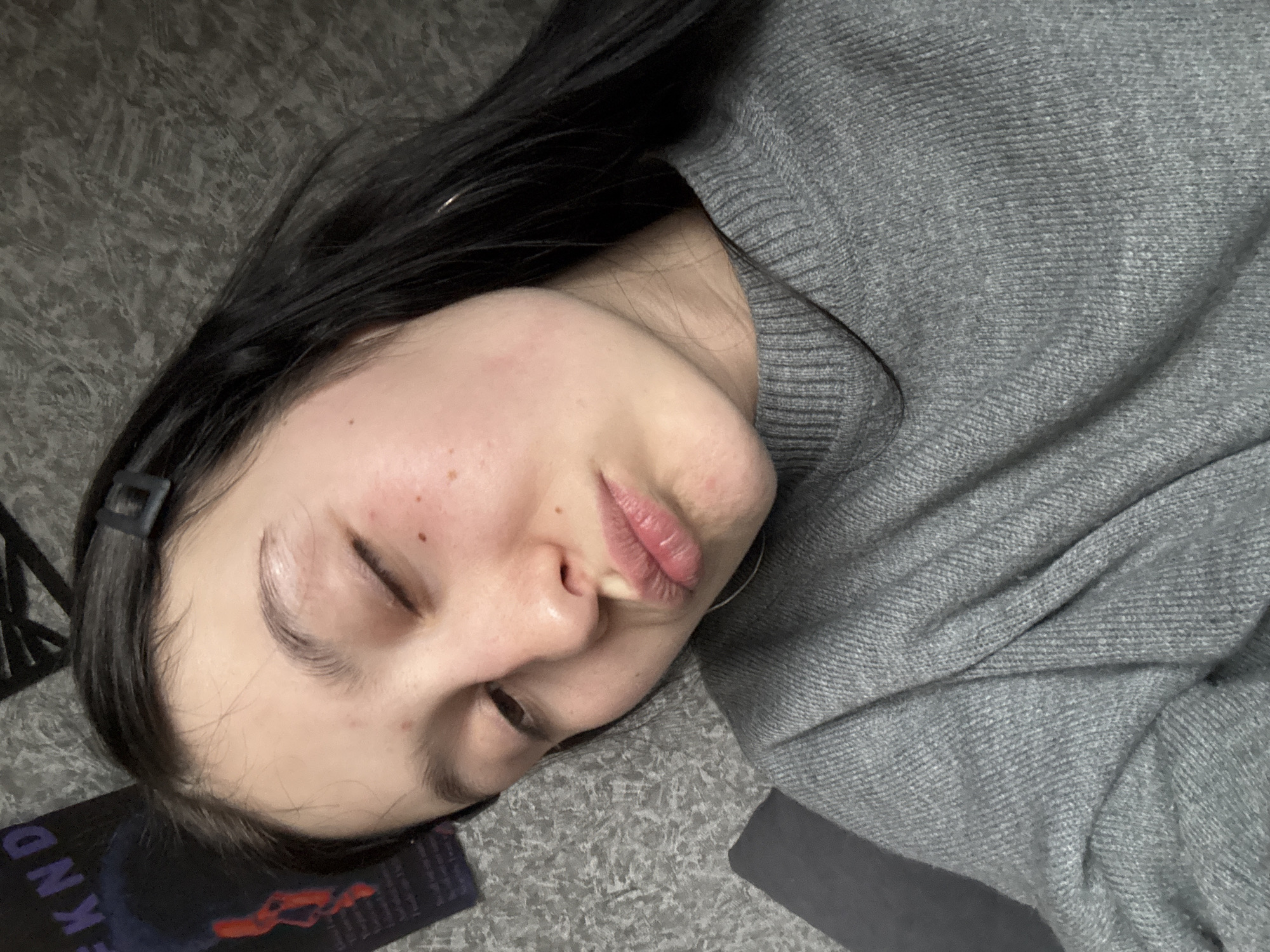 A young woman with dark hair lies on a gray carpet or rug, wearing a gray sweater. She is positioned at an angle, with her head tilted towards the lower left of the frame. Her eyes are mostly closed, and her expression is relaxed. A black headband is visible in her hair. A black book or album with the letters "EKND" partially visible rests near her head. The location, Фоки, Russia, cannot be determined from the visual information.