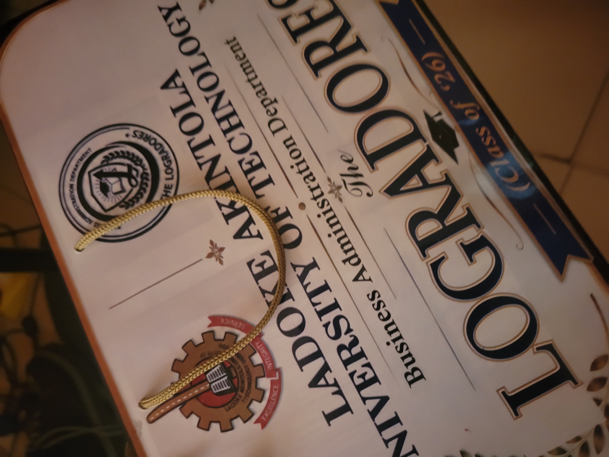 The image shows a close-up, angled view of a certificate or diploma. The document features text and graphics related to an academic institution. Prominently displayed are the words "LADOKE AKINTOLA UNIVERSITY OF TECHNOLOGY" and "Business Administration Department". Below this, the word "LOGRADORES" is emblazoned in large, bold letters, with "(Class of '26)" to its right. The certificate also includes various seals and decorative elements, such as a graduation cap graphic, a laurel wreath, and a gear-like emblem with text that appears to include "EXCELLENCE" and "INTEGRITY". The document is adorned with golden braided cords, suggesting it is a formal award. The lighting suggests indoor conditions, and the background is out of focus, hinting at a table or surface. The overall impression is that of an academic recognition, likely a graduation certificate. The location is unspecified.
