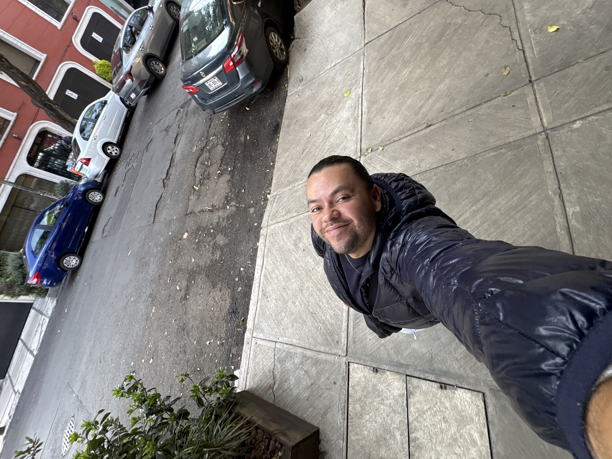 A medium-high angle shot features a man standing on a concrete sidewalk adjacent to a street with parked cars. The man is smiling at the camera, wearing a puffy navy blue jacket. Cars of varying colors (blue, white, silver, gray) are parked along the left side of the street, near a building with red brick walls. The street appears to be in a section of Mexico City, Mexico. A planter with greenery is visible in the lower left corner. The sidewalk is comprised of large square concrete tiles.