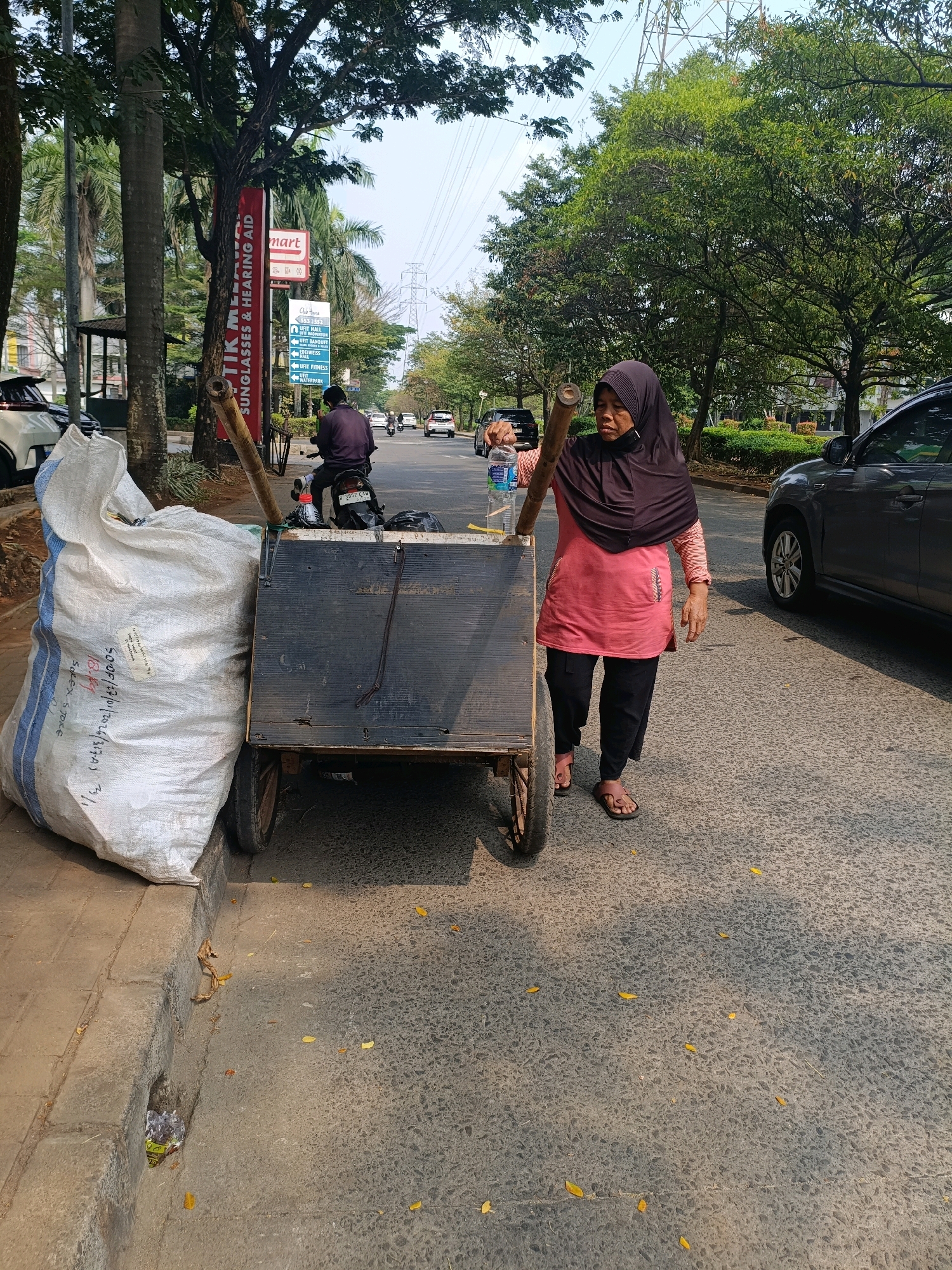 A woman wearing a dark hijab and a pink long-sleeved shirt is pushing a wheeled cart on a street in Tangerang, Indonesia. The cart is laden with a large white bag filled with what appears to be recyclables. The woman is walking on the right side of the street, next to the curb. A dark-colored car is parked on the right side of the road, and another dark-colored car is moving away from the camera on the left side of the road. Trees line the street, casting shadows on the pavement. A motorcycle with a rider is also visible in the background. A red banner with text in Indonesian hangs from a tree on the left side of the frame. The scene appears to be a typical street in a busy urban area.