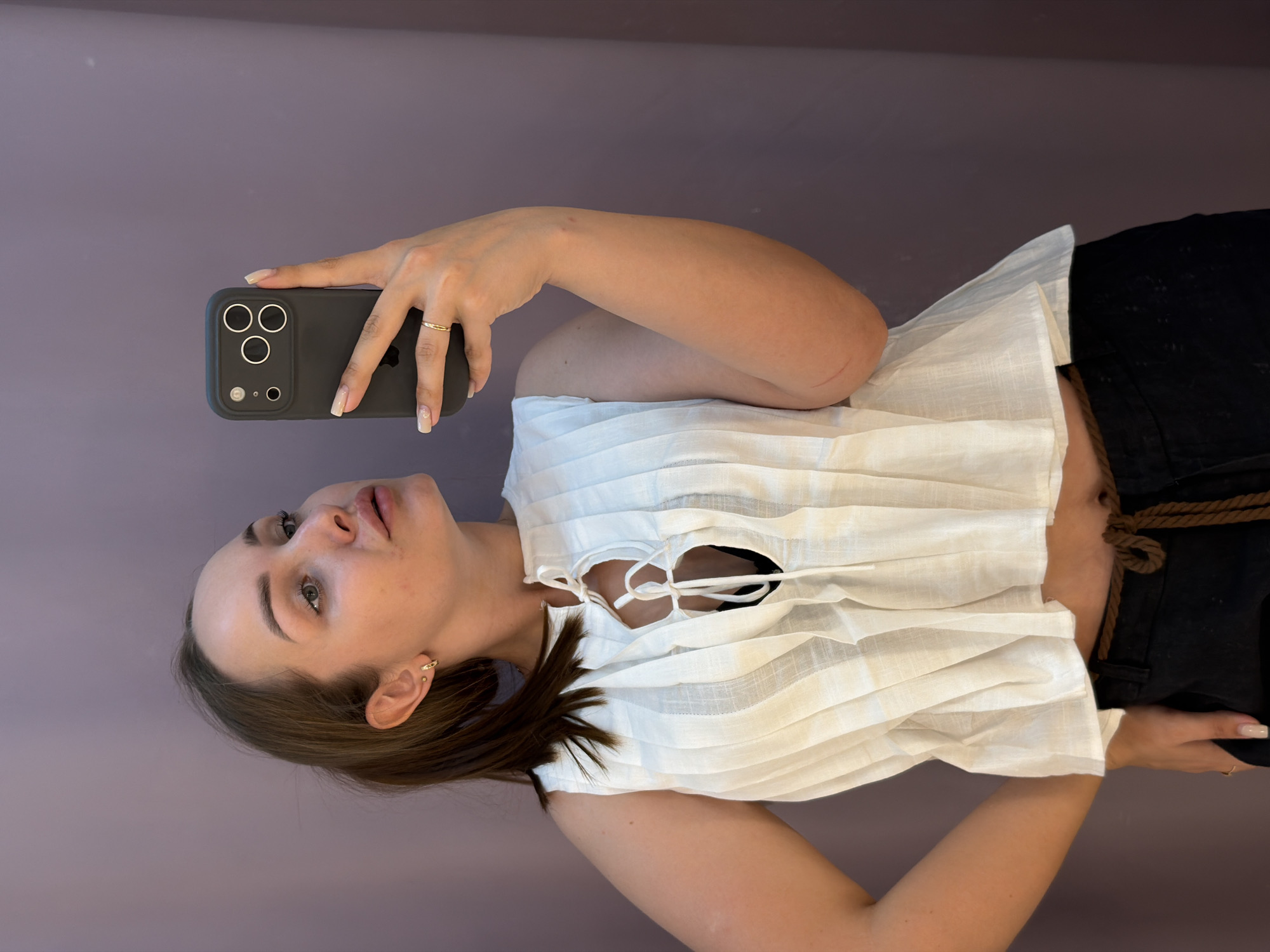 A young woman is taking a selfie. She is holding a gray phone with three rear-facing cameras. She is wearing a white blouse with ties at the front and black pants with a rope belt. She has light brown hair, fair skin, and a gold ring on her finger. The background is a plain lavender wall. It is unlikely for sure if this image was captured in Chaikovsky, Russia without additional information.