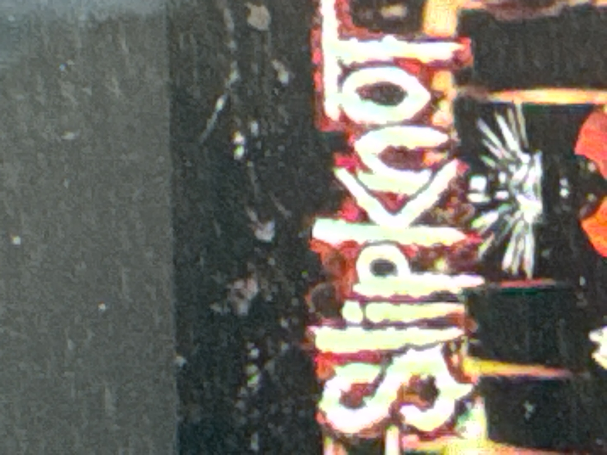 The image displays the word "Slipknot" in large, stylized, glowing white letters with a red outline, set against a dark, blurred background. Below the word, several dark, indistinct figures are visible, some wearing what appear to be black t-shirts with radiating line patterns. The overall scene is grainy and appears to be taken in a dimly lit environment, possibly a concert or event. No specific location in Kuta Selatan, Indonesia can be identified from the visual information.