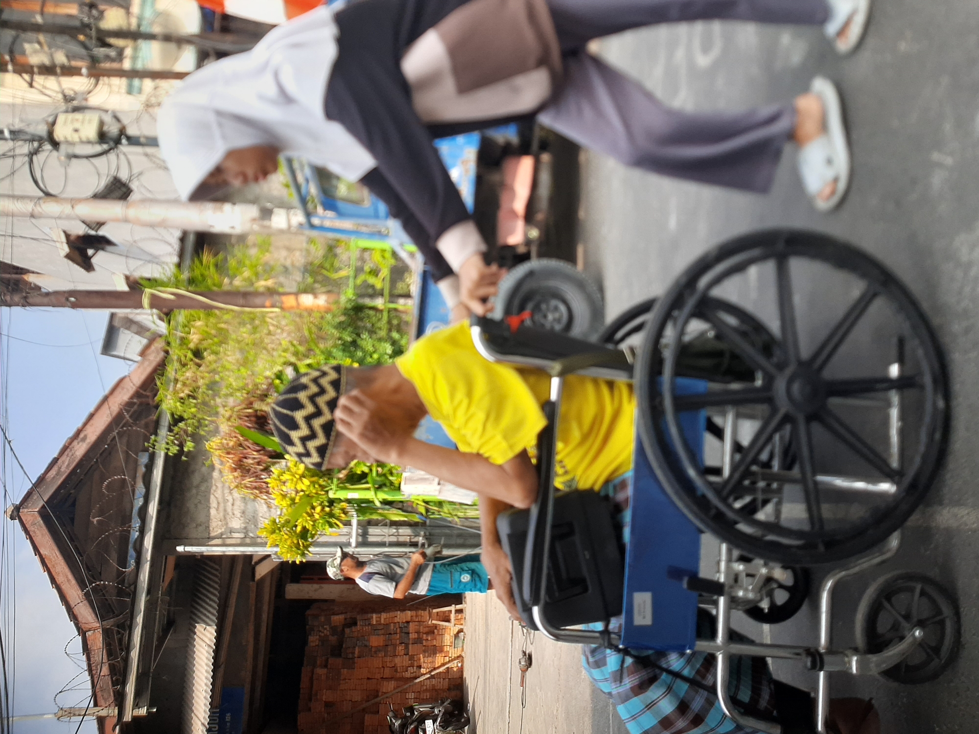 In Central Jakarta, Indonesia, a woman wearing a white hijab and a dark long-sleeved shirt with light brown accents pushes a man in a wheelchair. The man is wearing a yellow t-shirt and a black and gold patterned cap, with his hand on his face, appearing to be looking down. He is also covered by a blue and brown checkered cloth. The wheelchair is blue with black wheels. The woman is wearing gray pants and white sandals. In the background, a blue truck is visible, as well as a building with a corrugated metal roof and a pile of bricks. Another man in a gray t-shirt and blue shorts stands near the building, holding a tool. The scene appears to be on a street in an urban area.