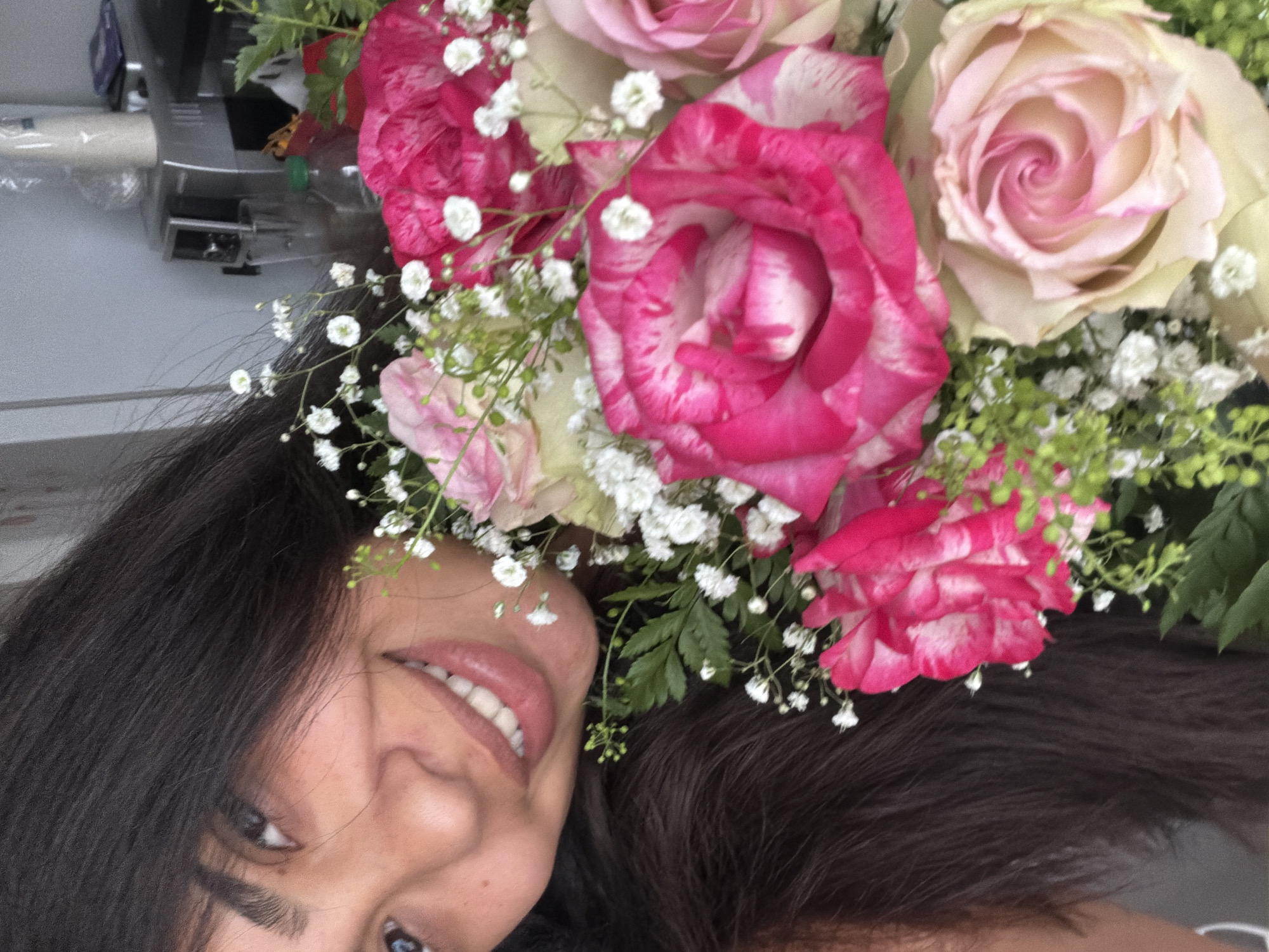 A close-up, selfie-style photograph shows a woman with long, dark hair smiling with a bouquet of pink and white roses. The woman is in the foreground, and the bouquet partially obscures her face. The background appears to be an indoor setting with some kitchen appliances visible. There are no visible landmarks to suggest a specific location within Duisburg, Germany. The scene is well-lit, with the focus on the woman and the flowers.