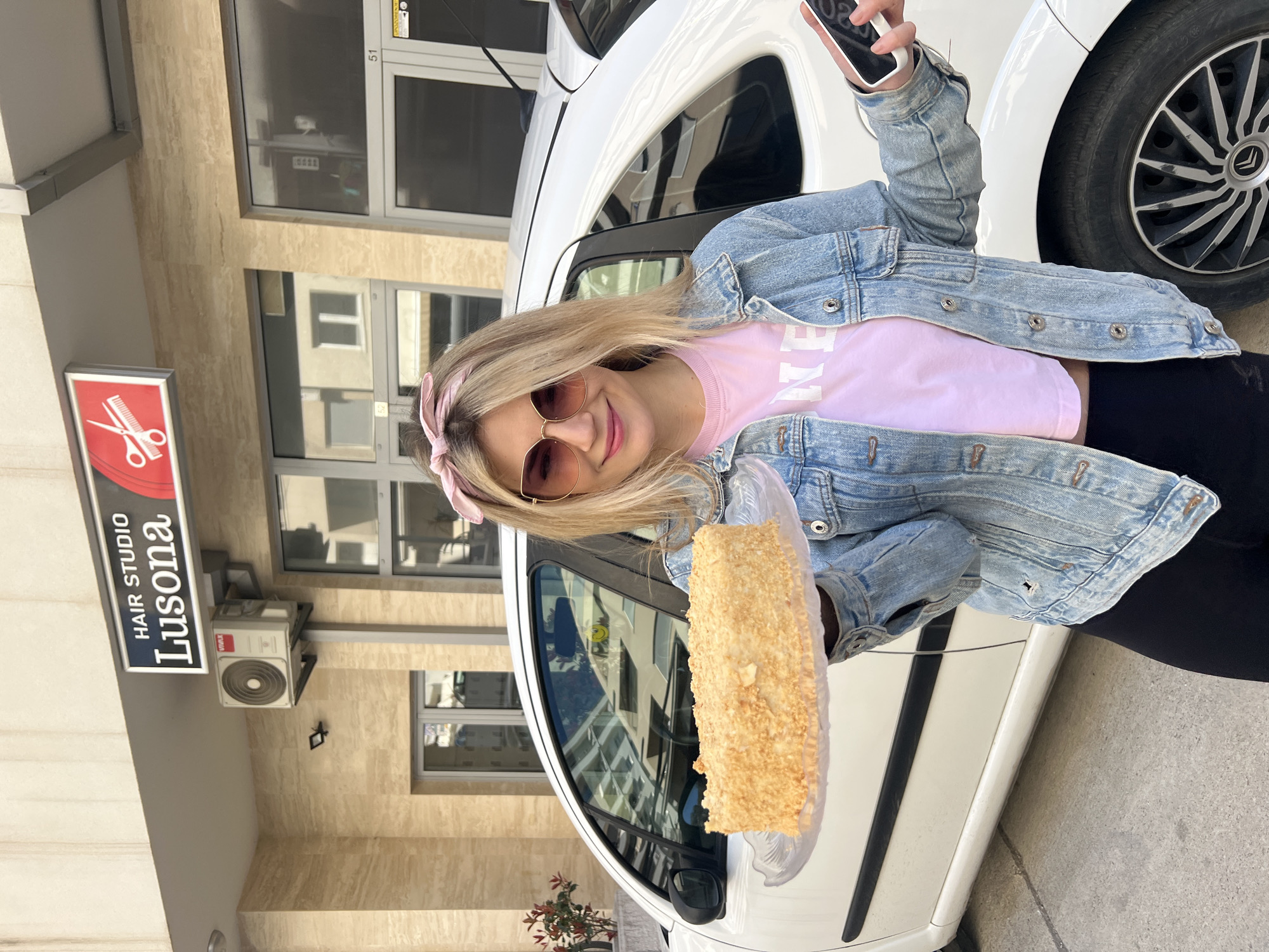 A woman is holding a large piece of cake in front of a white car. She is smiling and taking a selfie with a smartphone. The woman is wearing a denim jacket, a pink T-shirt, black leggings, and sunglasses, and has a pink headband in her blond hair. The car is parked outside a building identified by a sign as "HAIR STUDIO Lusona." An air conditioning unit is also visible on the building's facade. Based on the "Lusona" sign and architectural details, the location appears to be in the city of Budva, Montenegro.