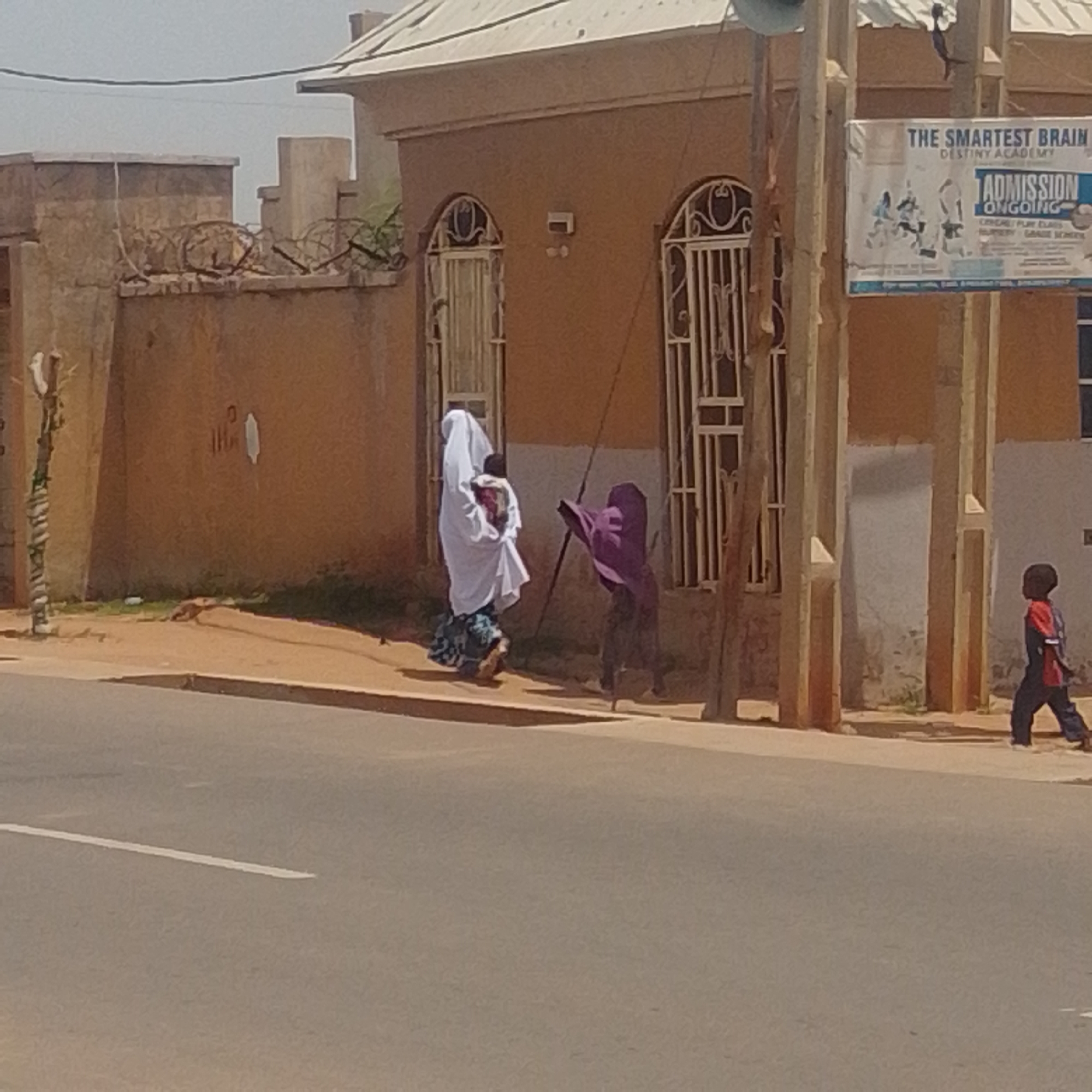 In Gombe, Nigeria, a street scene unfolds in front of a building with ornate metal-barred windows and a light-colored roof. A man, possibly a child, wearing a blue and red outfit, walks on the right side of the frame away from the camera, facing the street. To the left, a woman wearing a white hijab and a patterned skirt carries a child. Another figure, partially obscured, wears a dark purple garment and appears to be walking away from the woman. A promotional banner for "The Smartest Brain Destiny Academy" hangs on a pole to the right of the building, advertising admissions. The ground is a mix of dirt and asphalt, with a white line marking the edge of the road. The environment is overcast with a dry, dusty appearance.