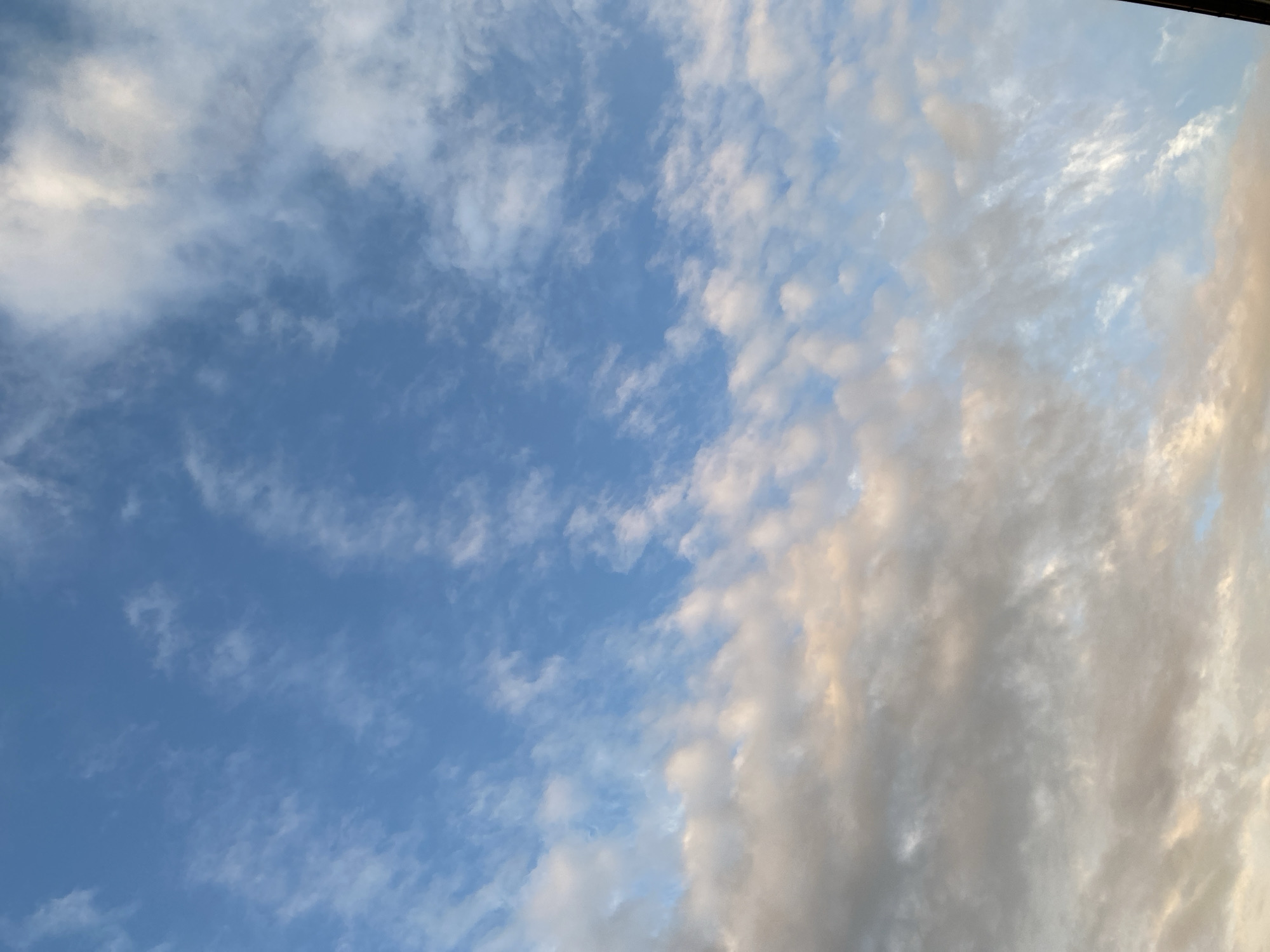 An image of the sky shows a mixture of white and gray clouds against a blue sky. The clouds are scattered across the frame, with some appearing wispy and others more dense. The lighting suggests it may be late afternoon or early morning, as some of the clouds have a warm, golden hue. The perspective is from a ground-level view looking upwards. The image does not contain any discernible elements that would indicate a specific city or location.