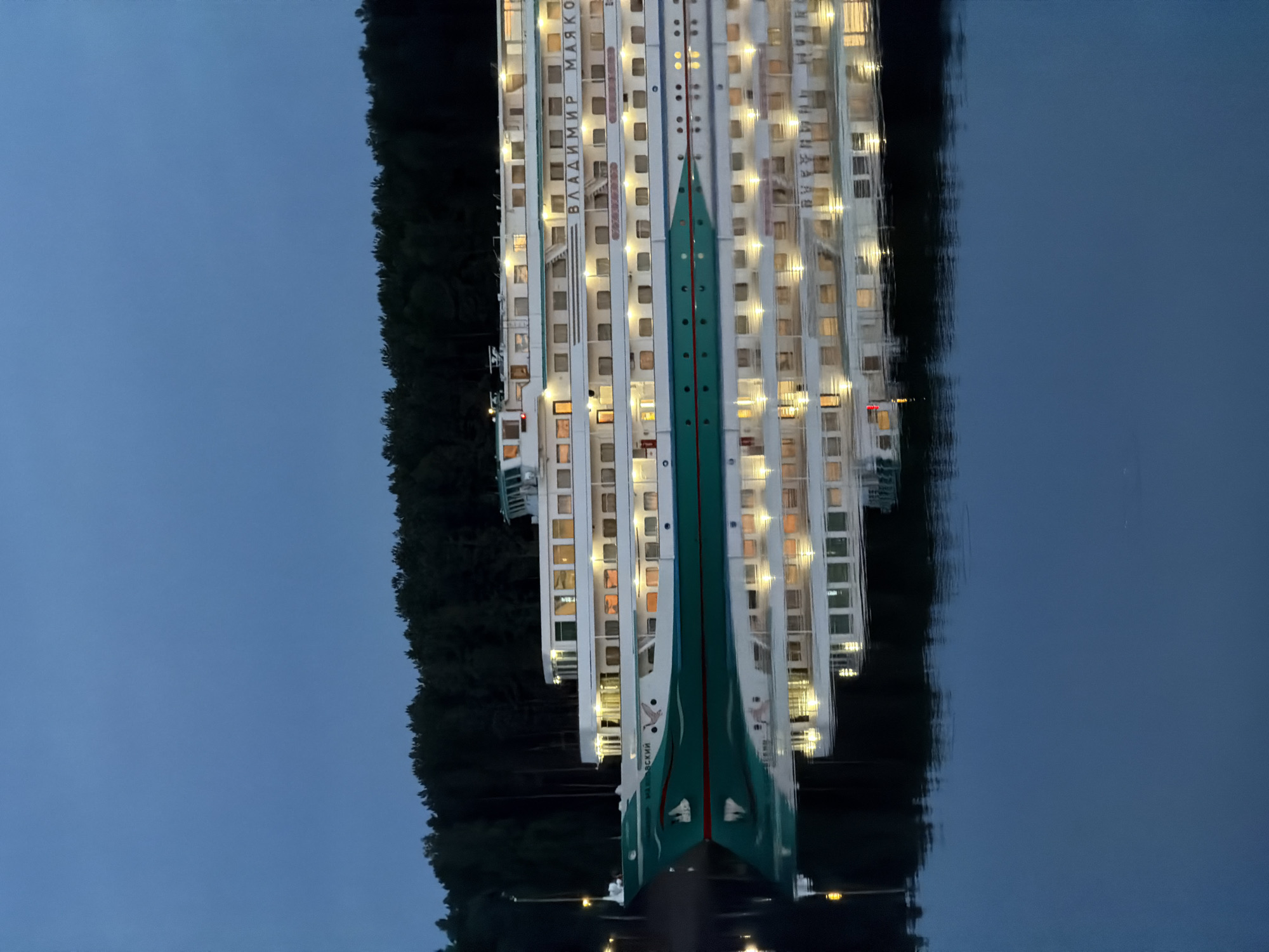 The image shows a cruise ship reflected in water at night. The ship is lit up with many windows showing light. The name "ВЛАДИМИР МАЯКОВСКИЙ" is visible on the side of the ship. There is dark vegetation on the left side of the ship. The image is a reflection in the water. It shows a scene near the city of Chaikovsky, Russia.