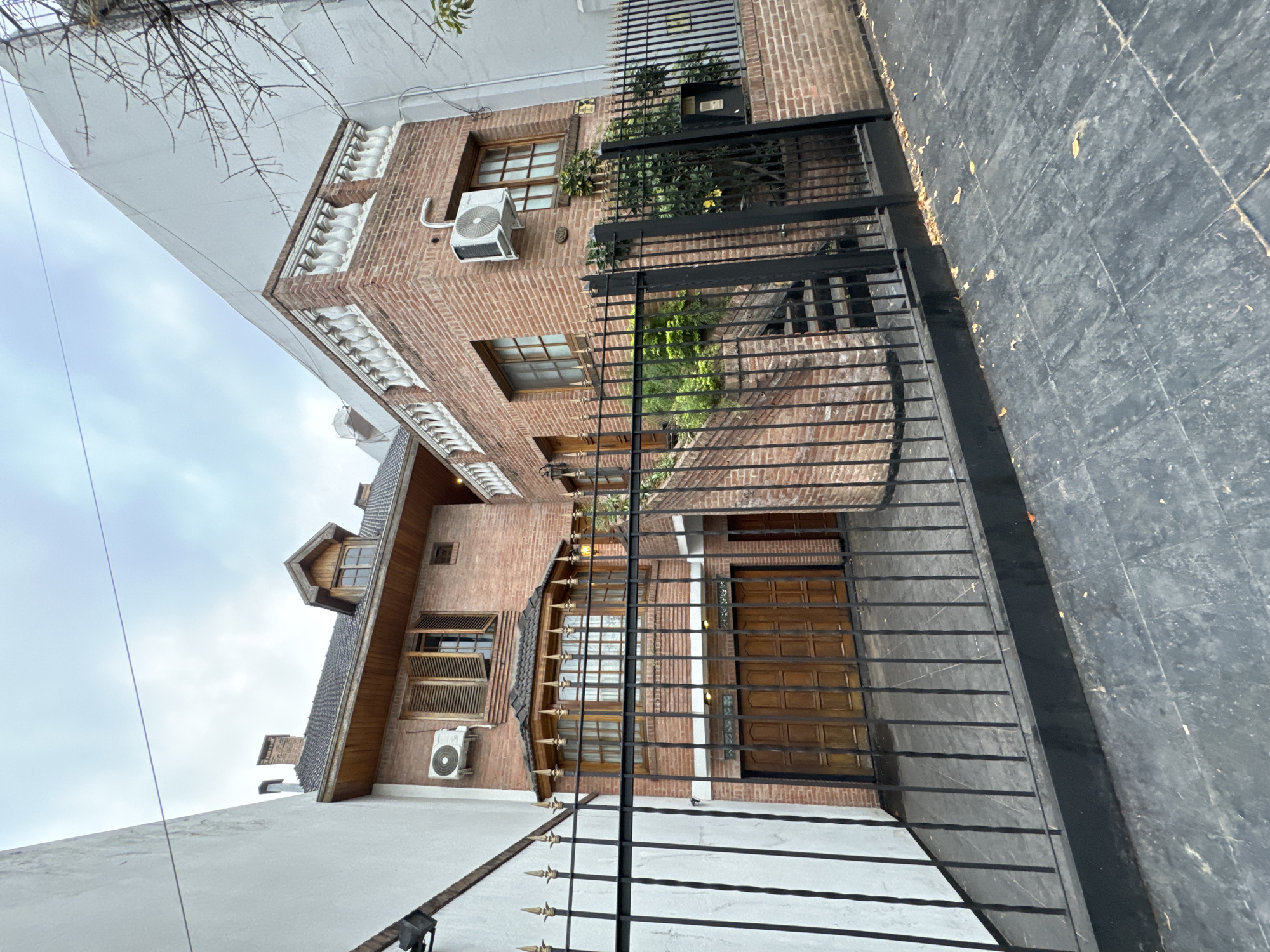 The image depicts a residential street scene in Buenos Aires, Argentina. The perspective is tilted, showing two adjacent brick buildings behind a black wrought-iron fence. The building on the right is two stories with an air conditioning unit visible on the exterior wall. A black box is mounted near the top of the wall. The building on the left is a single-story, white-walled structure, and a two-story brick building with a wood framed roof sits behind the white building. The sidewalk is tiled with dark gray slabs. Bare branches of a tree are visible in the top left corner. No people are visible in the shot.