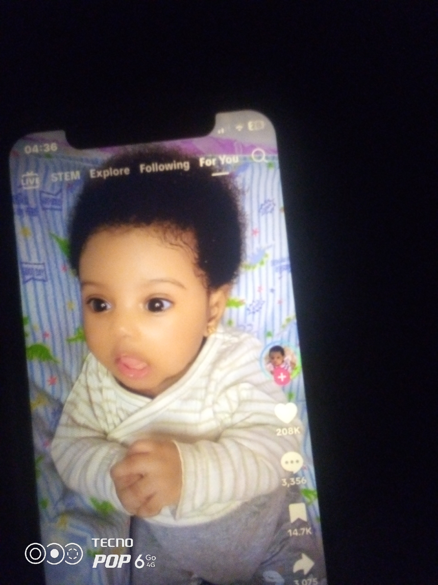 The image shows a close-up of a baby's face and upper body, viewed on a smartphone screen. The baby, who appears to be of African descent, has dark curly hair, brown eyes, and is wearing a white and gray striped shirt. The baby's tongue is slightly sticking out, and their hands are clasped in front of them. The background behind the baby on the phone screen appears to be a light blue and white striped crib with some cartoon dinosaur graphics.
Overlaid on the phone screen are various user interface elements typical of a social media app. These include a timestamp "04:36", navigation options like "LIVE", "STEM", "Explore", "Following", and "For You", a search icon, a small circular profile picture with a plus sign, a heart icon with "208K" likes, a speech bubble icon with "3,356" comments, a bookmark icon with "14.7K" saves, and an arrow icon with "3,075" shares. In the bottom left corner of the phone screen, there is a logo that reads "TECNO POP 6 4G".
The image is taken from an angle, and the smartphone is placed on a dark surface, likely a table or a lap. The lighting is dim, casting some shadows. The overall scene is a photograph of a smartphone displaying a video or image of a baby, with the phone's interface visible. The location in the city of Howayi, Nigeria is not discernible from the image itself, as it only shows the content displayed on a phone screen and the immediate surroundings of the phone.