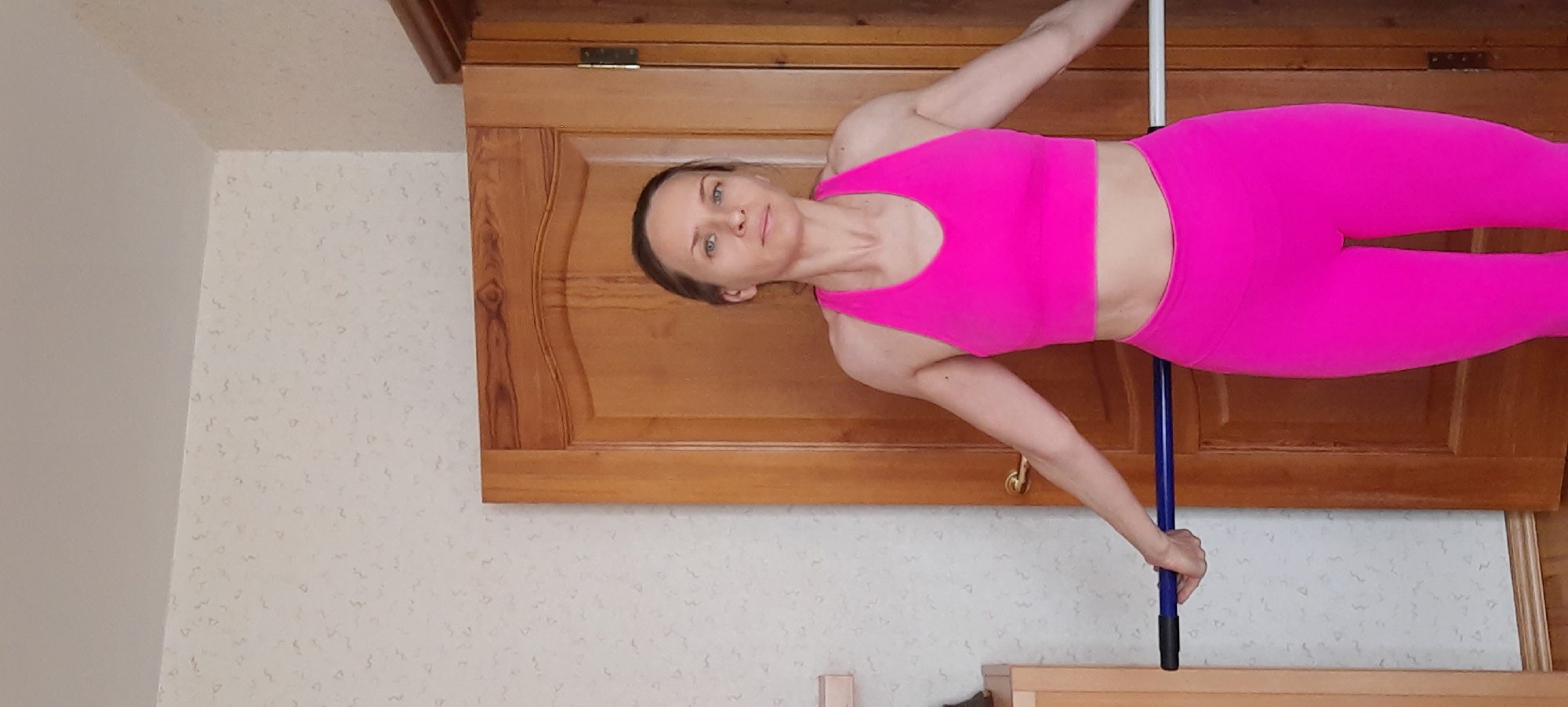 An adult woman is depicted indoors, wearing a vibrant hot pink athletic sports bra and matching leggings. She has light skin, and her light-colored hair is pulled back, framing a calm and focused expression as she looks directly towards the viewer. The woman is actively holding a blue and black pole or stick horizontally across her midsection, gripping it firmly with both hands. Her body is oriented horizontally within the frame, indicating she is performing a static strength or balance exercise.
The background primarily features a large, light brown wooden cabinet or wardrobe, distinguished by carved panels and visible hinges at the top. Adjacent to and below the wooden furniture, a wall with light-colored, subtly patterned wallpaper is visible. The overall setting suggests a domestic environment, possibly a home in Moscow, Russia. The lighting indicates it is daytime. There is no visible text in the image.