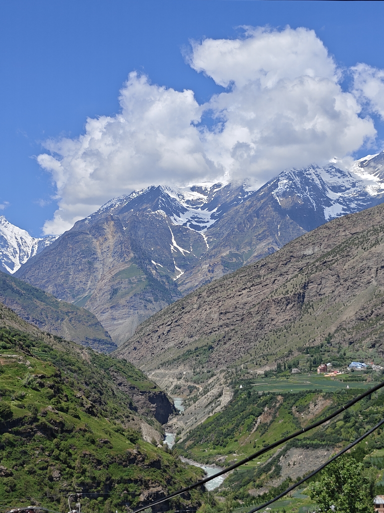 This is a vertical, eye-level, full shot of the mountainous landscape surrounding Keylang, India. In the foreground, a steep, verdant slope is covered in lush green vegetation and trees. A river flows through the valley below, with a small village nestled on a green plateau in the mid-ground. The village consists of several buildings, some with colorful roofs, and agricultural fields. The background is dominated by imposing, rocky mountains, some of which are snow-capped. The sky is a vibrant blue, with large, fluffy white clouds scattered across it, partially obscuring the mountain peaks. There are no people or animals visible in the image, nor any explicit actions or interactions. The scene conveys a sense of natural beauty and the serene environment of a remote mountain region.