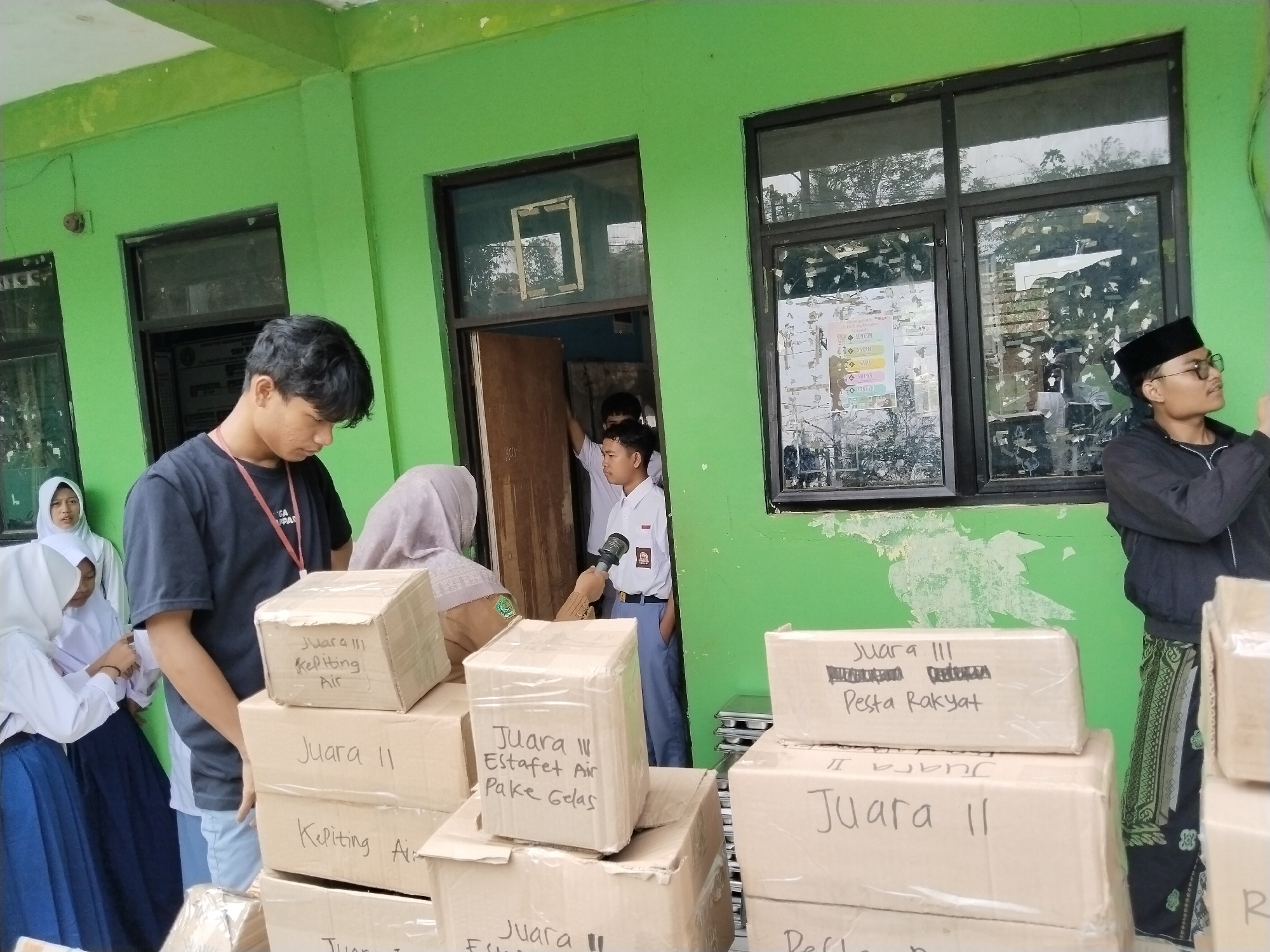 The image depicts an outdoor scene in front of a green-painted building, likely a school in Serang, Indonesia. A group of individuals, some wearing school uniforms and others in casual attire, are gathered around several cardboard boxes. The boxes are stacked and some have handwritten labels in Indonesian, such as "Juara III" (3rd Place) and "Pesta Rakyat" (People's Party).
In the foreground, on the left, two young women in white school uniforms and headscarves are attending to something near a stack of boxes. A young man wearing a black t-shirt stands behind them, his right arm visible. To the right, a man wearing glasses and a black peci hat is looking to the right, seemingly observing something. Another young man in a white school uniform stands in a doorway in the background. The environment suggests an event or distribution of goods, possibly awards or supplies, taking place in a semi-public area. The building's exterior shows signs of wear, with chipped green paint. The presence of windows with reflective glass suggests they are looking into an indoor space.