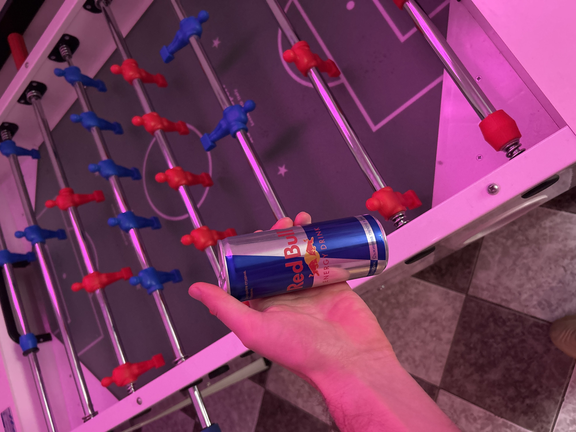 A person's hand holds a blue and silver Red Bull energy drink can over a foosball table. The table has rows of blue and red foosball players. The table is lit with pink light. The background suggests a tiled floor. The location is unknown and cannot be determined from the image. The city of Sofia, Bulgaria, cannot be confirmed based on the provided image.