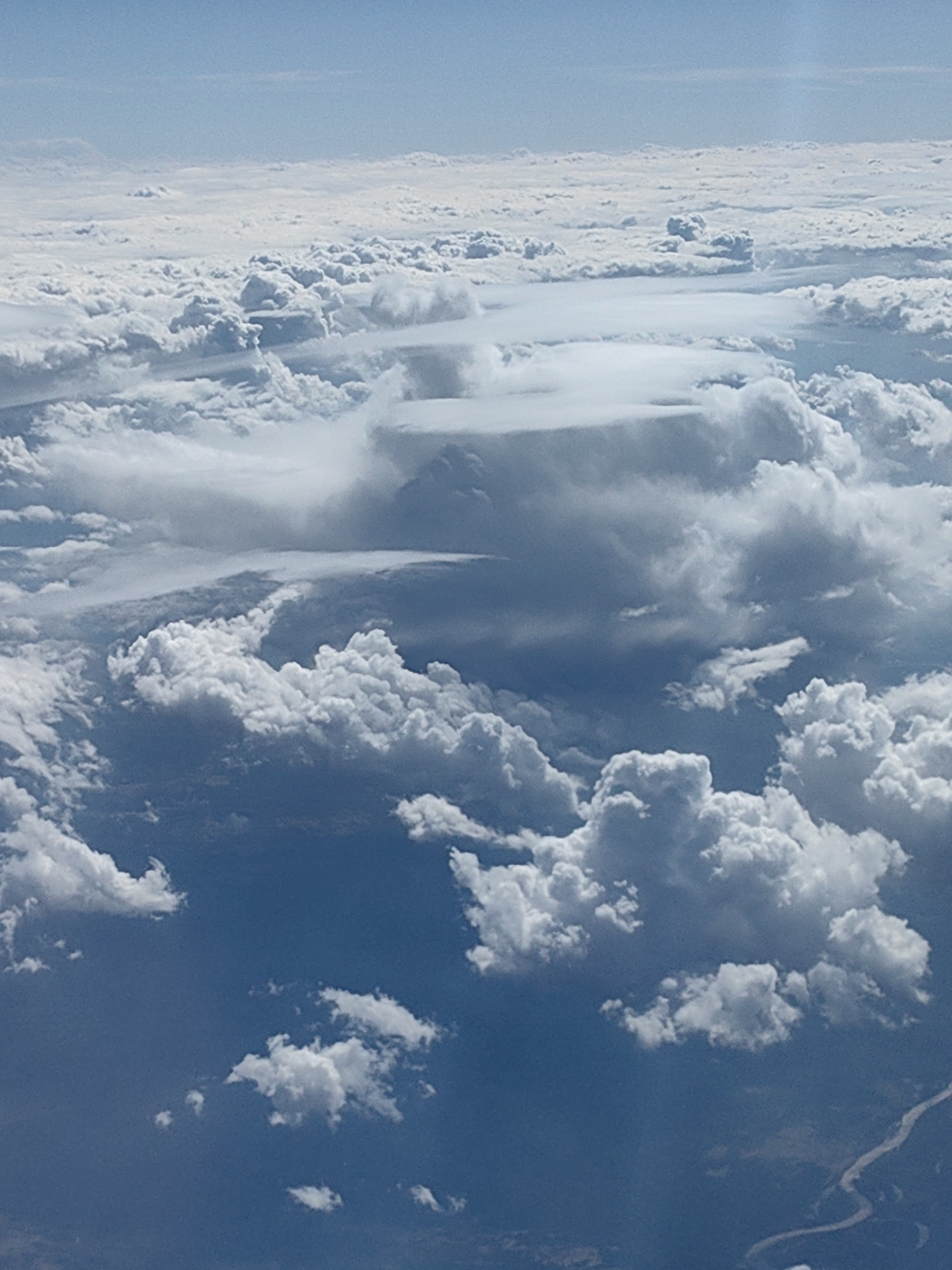 The image shows a view from above the clouds. The sky is visible at the top, transitioning into thick layers of white clouds with varying depths and textures. Beneath the clouds, glimpses of the land can be seen, including a river winding through the terrain. The location is potentially near Khimki, Russia, evidenced by the landscape visible through the clouds. There are no people, identifiable subjects, or distinct actions visible. The environment consists of the sky, cloud formations, and glimpses of the terrain below.