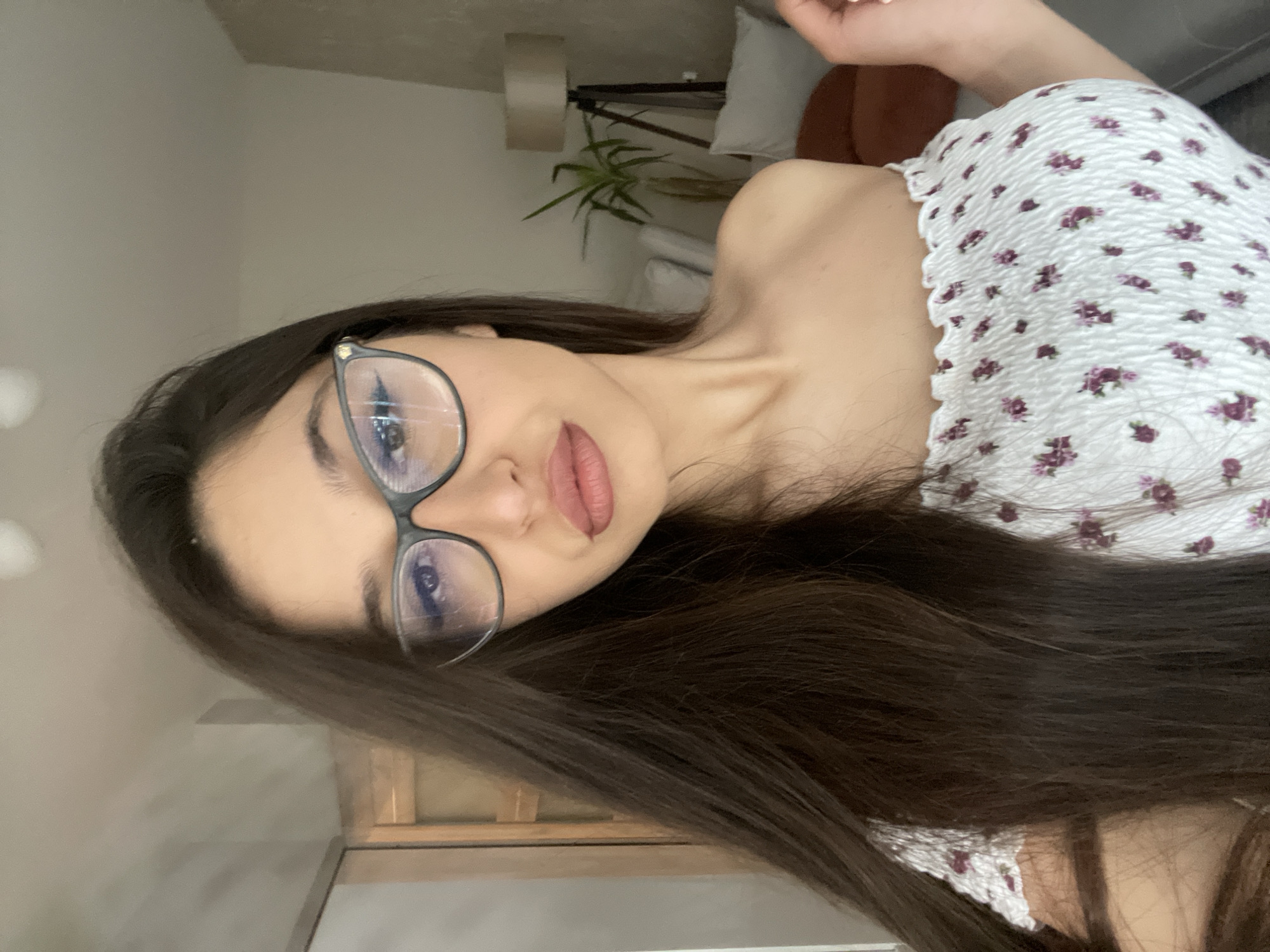 This is a portrait of a young woman wearing glasses and a floral-patterned top. She has long, dark brown hair that falls over her shoulders. Her eyes are visible through her glasses, and she is wearing lip gloss. The woman is looking towards the camera. The background is softly blurred, suggesting an indoor setting, possibly a room in a house. There are hints of furniture, including a lamp and some greenery, suggesting a domestic environment. The lighting appears to be natural or soft indoor lighting, and the time of day is indeterminate. The overall mood of the image is casual and relaxed. There is no visible text in the image.