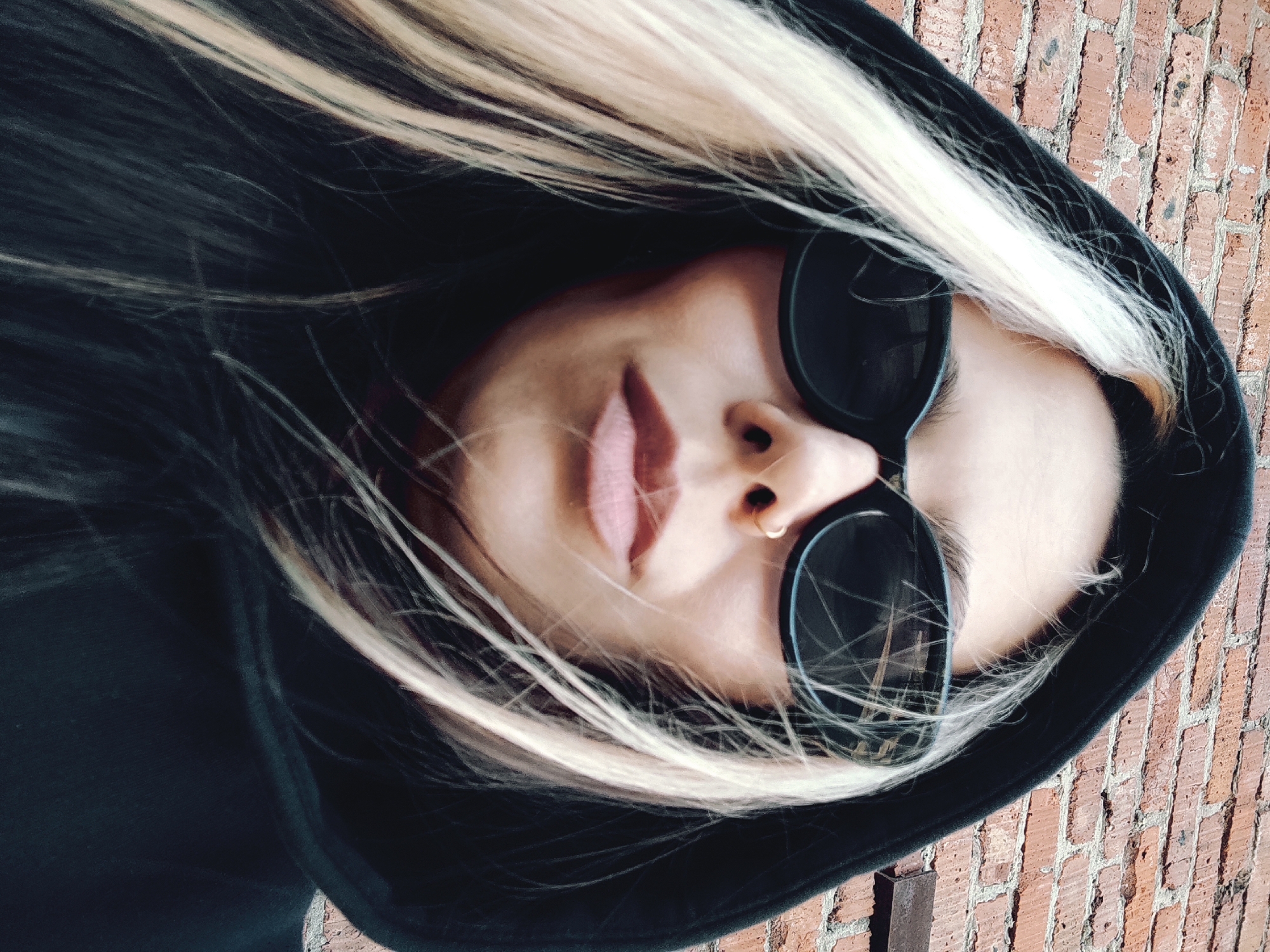 A person wearing sunglasses and a black hooded sweatshirt is facing the camera. The person has blonde and dark hair, and a nose piercing. The background is a brick wall. The scene appears to be outdoors. The location is not discernible to Chaikovsky, Russia.