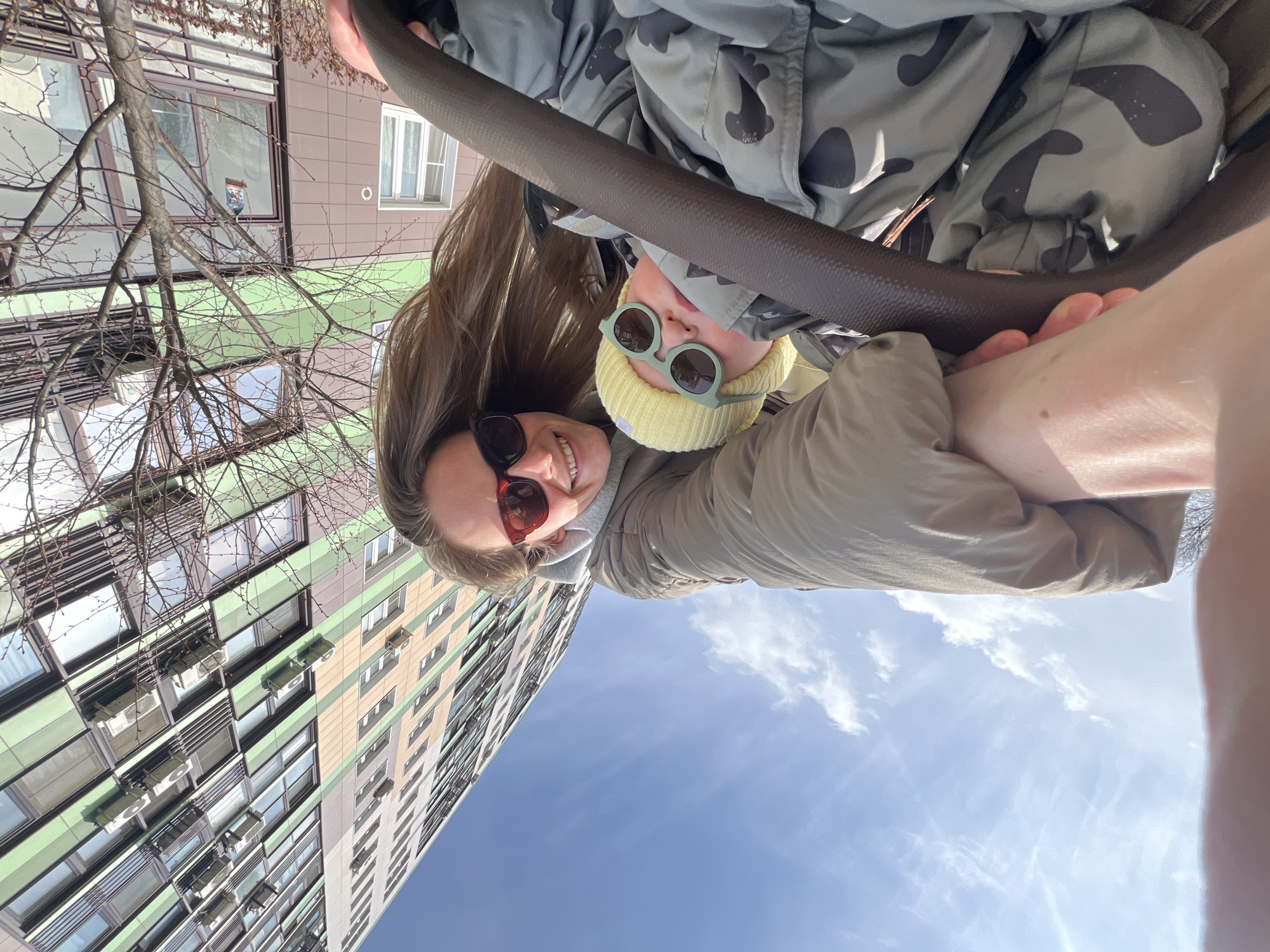Here is a forensic description of the image:
Two individuals are visible, likely a parent and child, possibly a mother and child, suspended upside down from a dark, possibly metal or plastic, cylindrical bar. The adult is wearing sunglasses with red lenses and a grey jacket. The child is wearing a yellow beanie and round, light-green sunglasses. A large building is visible in the background, with a multi-colored facade of light-green, beige and grey vertical panels and numerous windows, each adorned with a white air conditioning unit. The sky is blue with wispy white clouds. Bare tree branches appear on the left side of the frame, positioned against the building. The location is the city of Khimki, Russia. The individuals seem to be engaged in play or recreation at an outdoor playground. The camera angle is low, looking upward. The picture has been rotated.